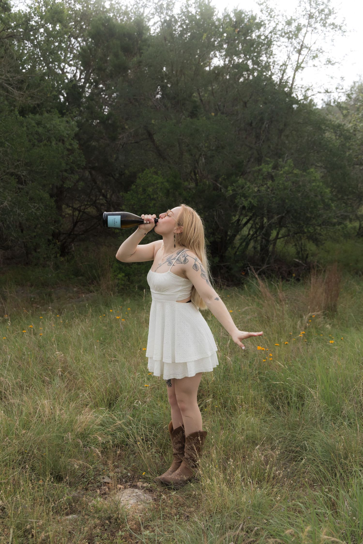 Blonde woman in white dress drinking from a wine bottle in a wildflower meadow.