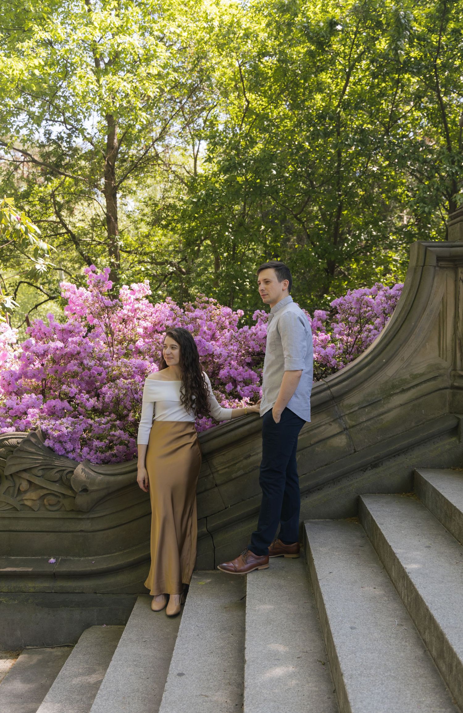 Couple posing on stone steps surrounded by blooming pink rhododendrons and lush green trees in a park.
