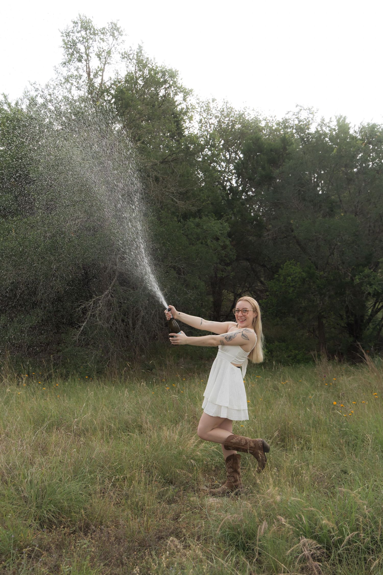 Blonde woman in white dress and cowboy boots spraying champagne in a green field.