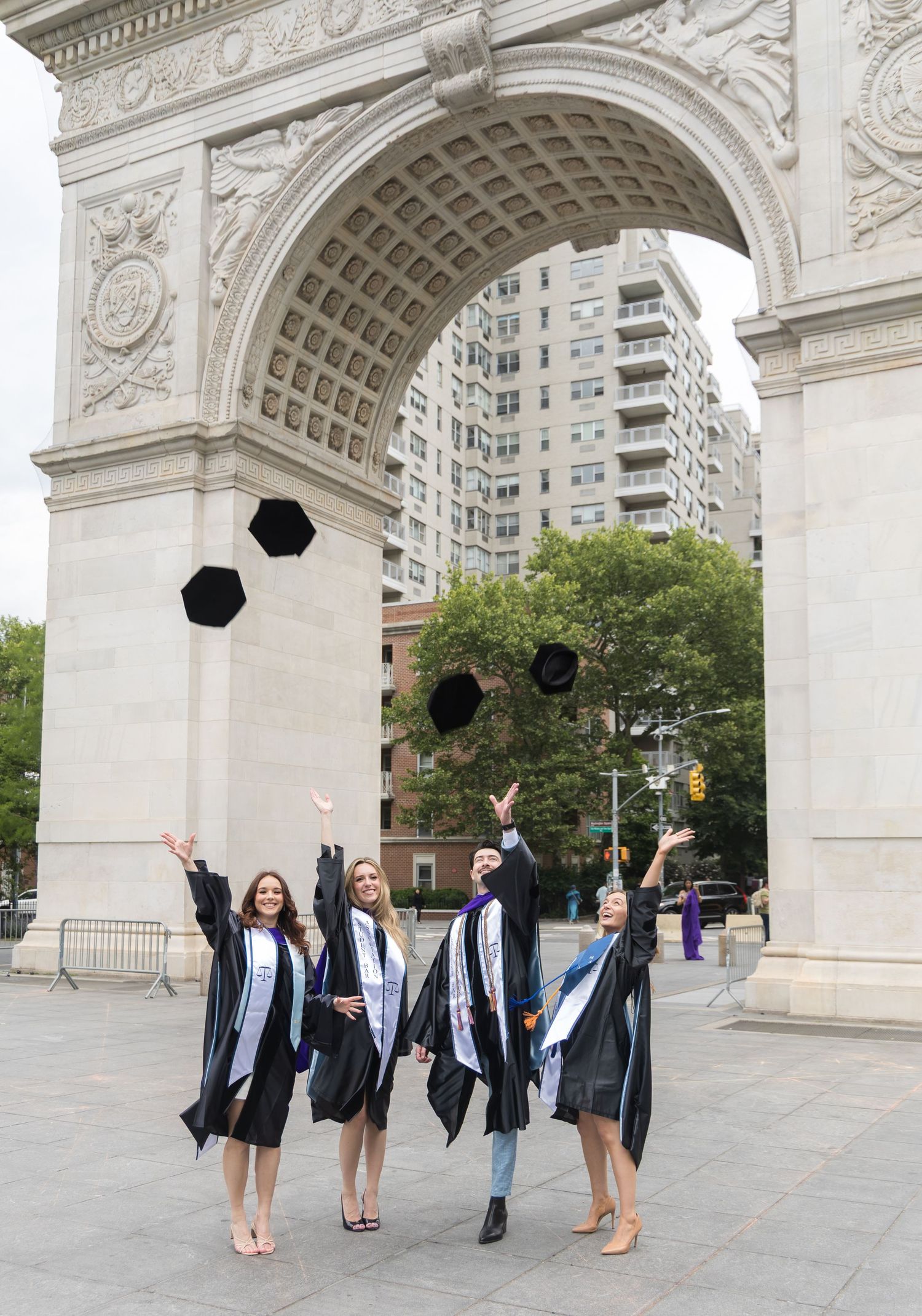Four graduates toss caps in air under Washington Square Park arch in New York City.