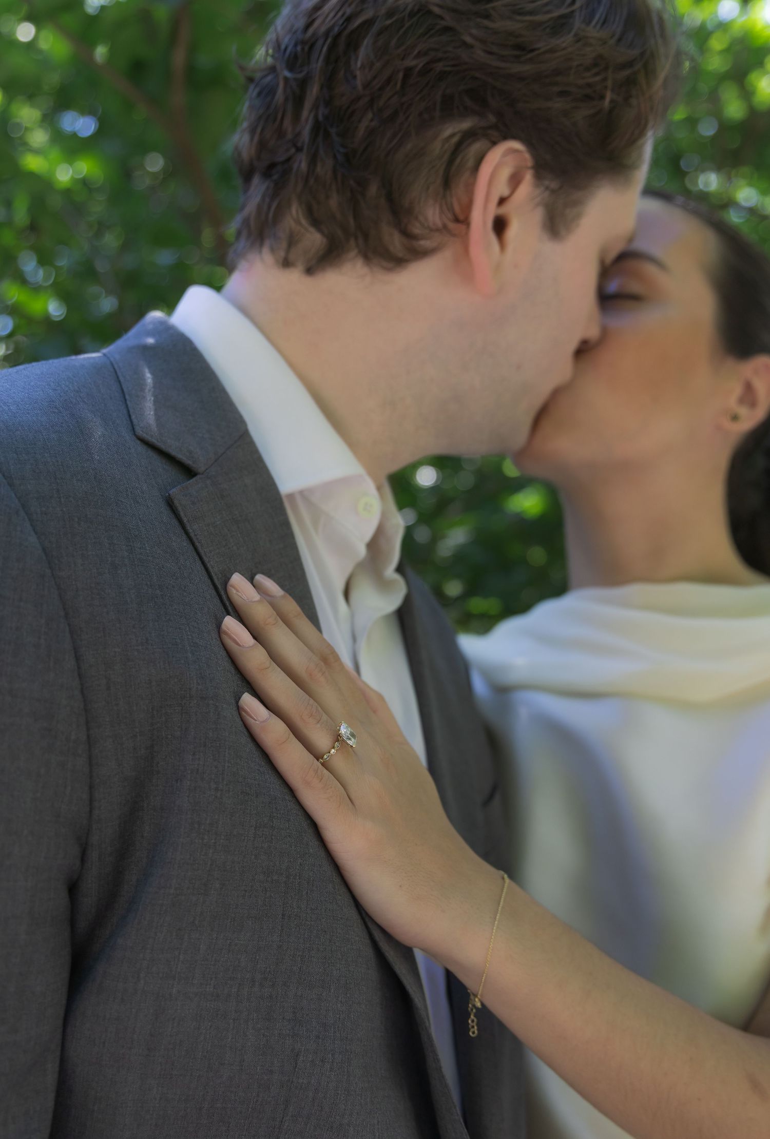 Couple sharing a romantic kiss, bride wearing diamond engagement ring, groom in gray suit, outdoor wedding ceremony.