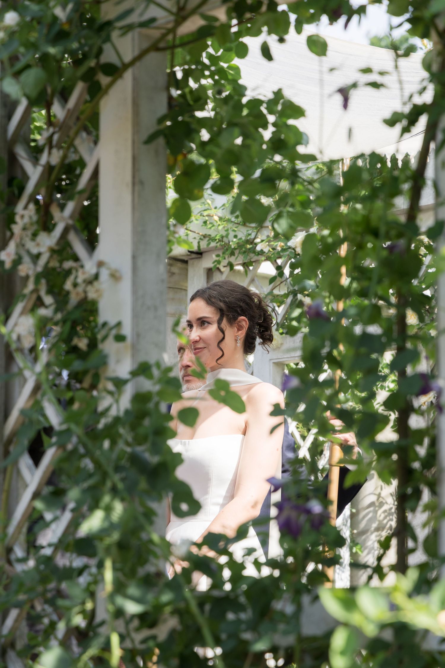 Bride in white dress standing among lush green garden foliage, partially framed by climbing vines and lattice.