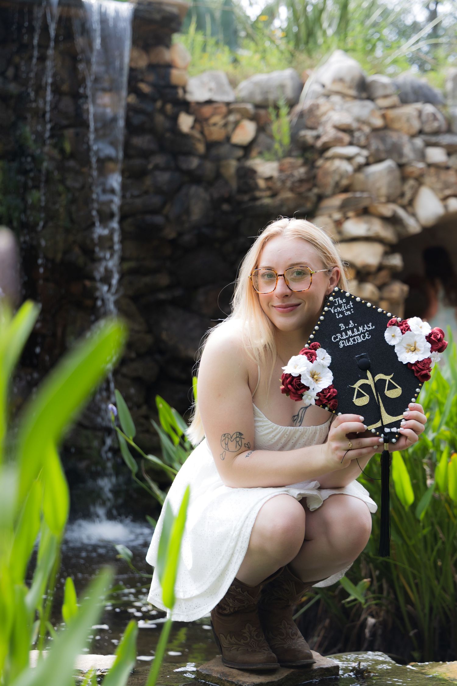 Blonde woman in white dress crouching by waterfall holding decorated graduation cap with flowers.