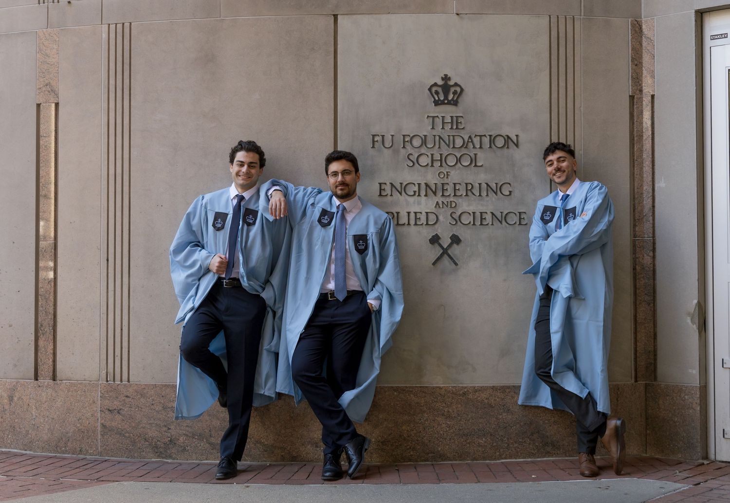 Three graduates in blue robes pose proudly outside the EU Foundation School of Engineering and Applied Science.