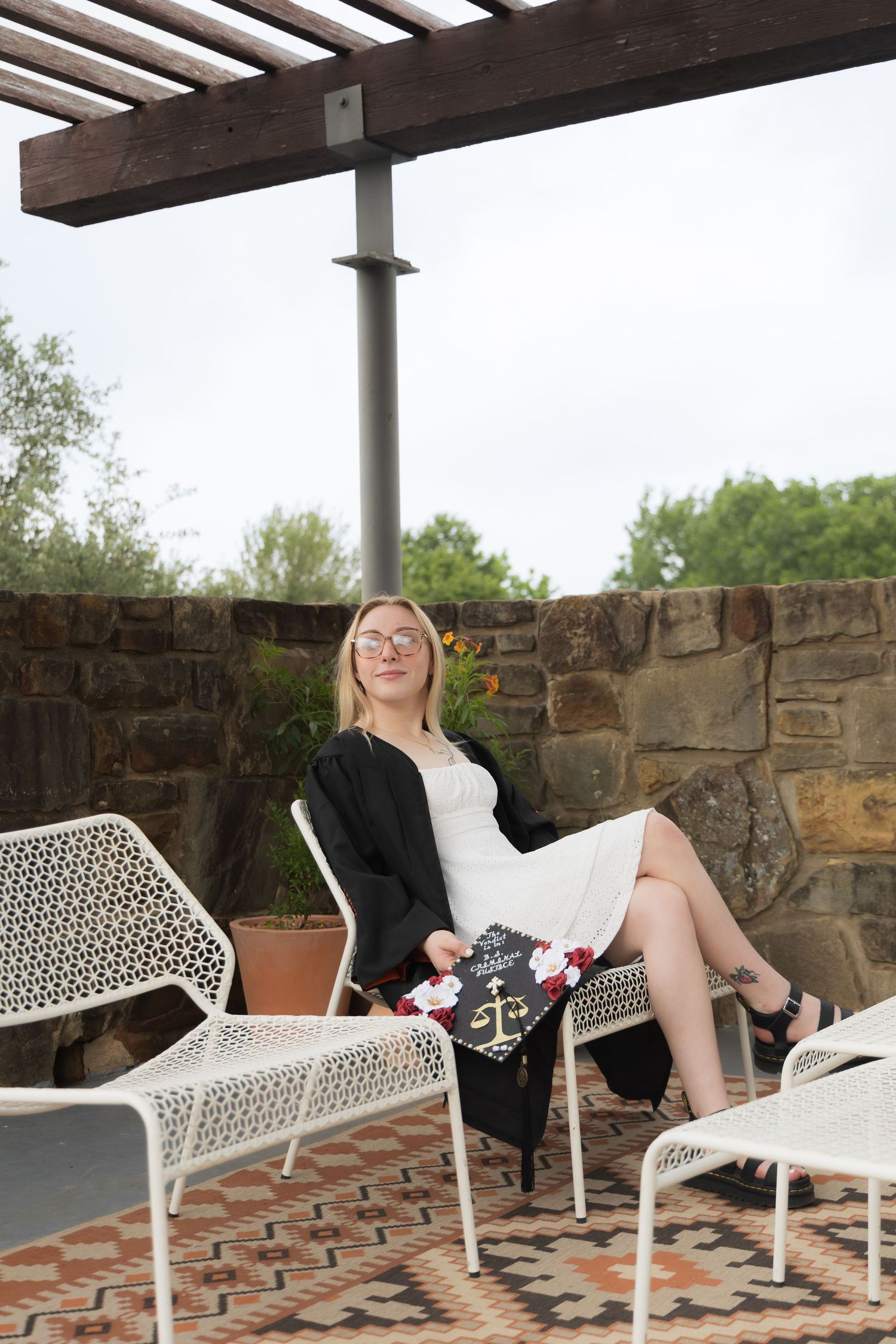 Blonde graduate in black gown sitting in white chair on stone patio holding decorated cap.