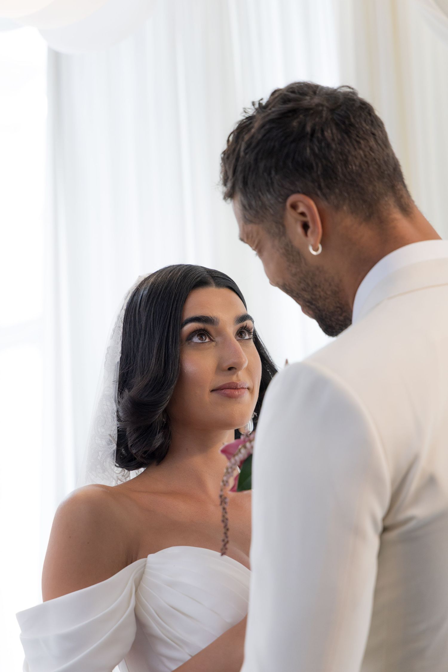 Romantic wedding couple shares an intimate moment against white curtains in elegant attire.