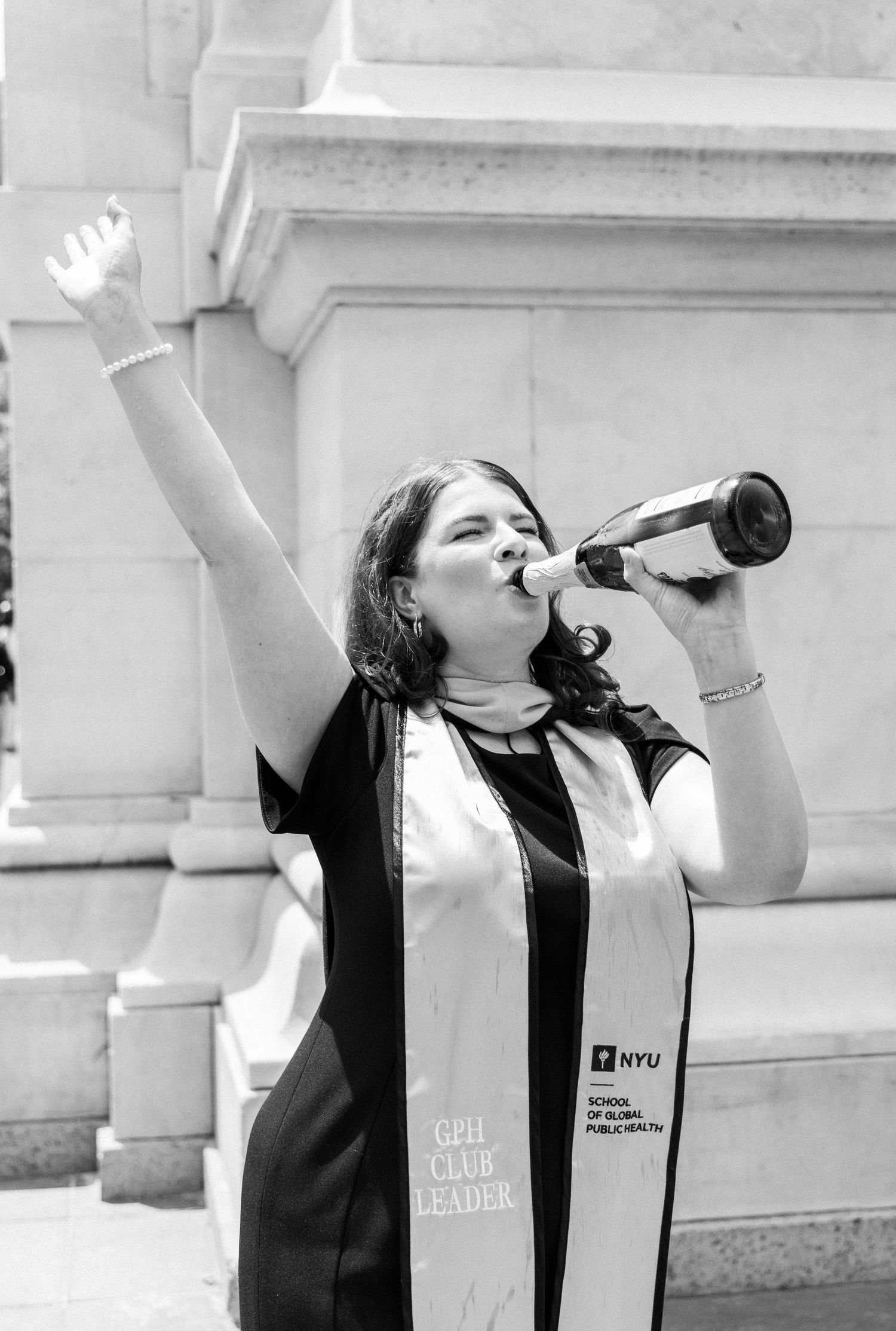 NYU School of Global Public Health graduate celebrating outdoors, raising arm and drinking from bottle in graduation regalia.