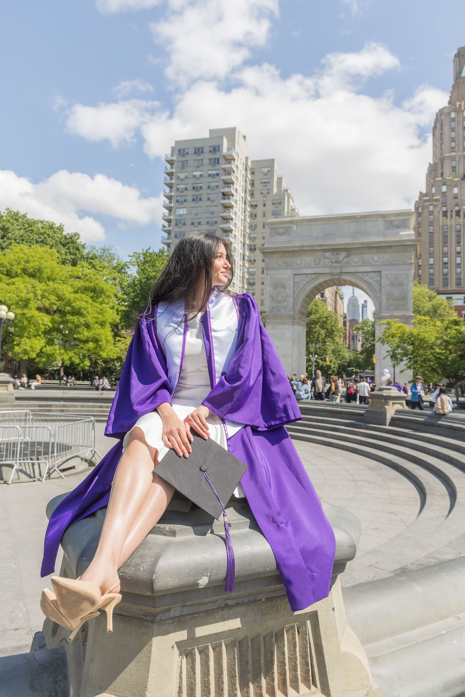 Graduate in purple cap and gown sits near Washington Square Park arch in NYC on a sunny day.