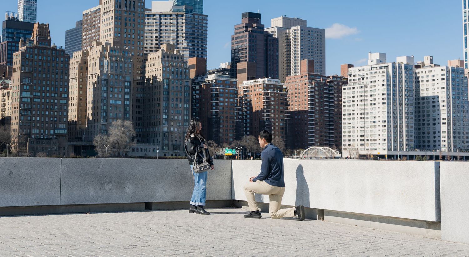 Man proposing to woman on rooftop terrace with Manhattan skyline skyscrapers in background on sunny day.