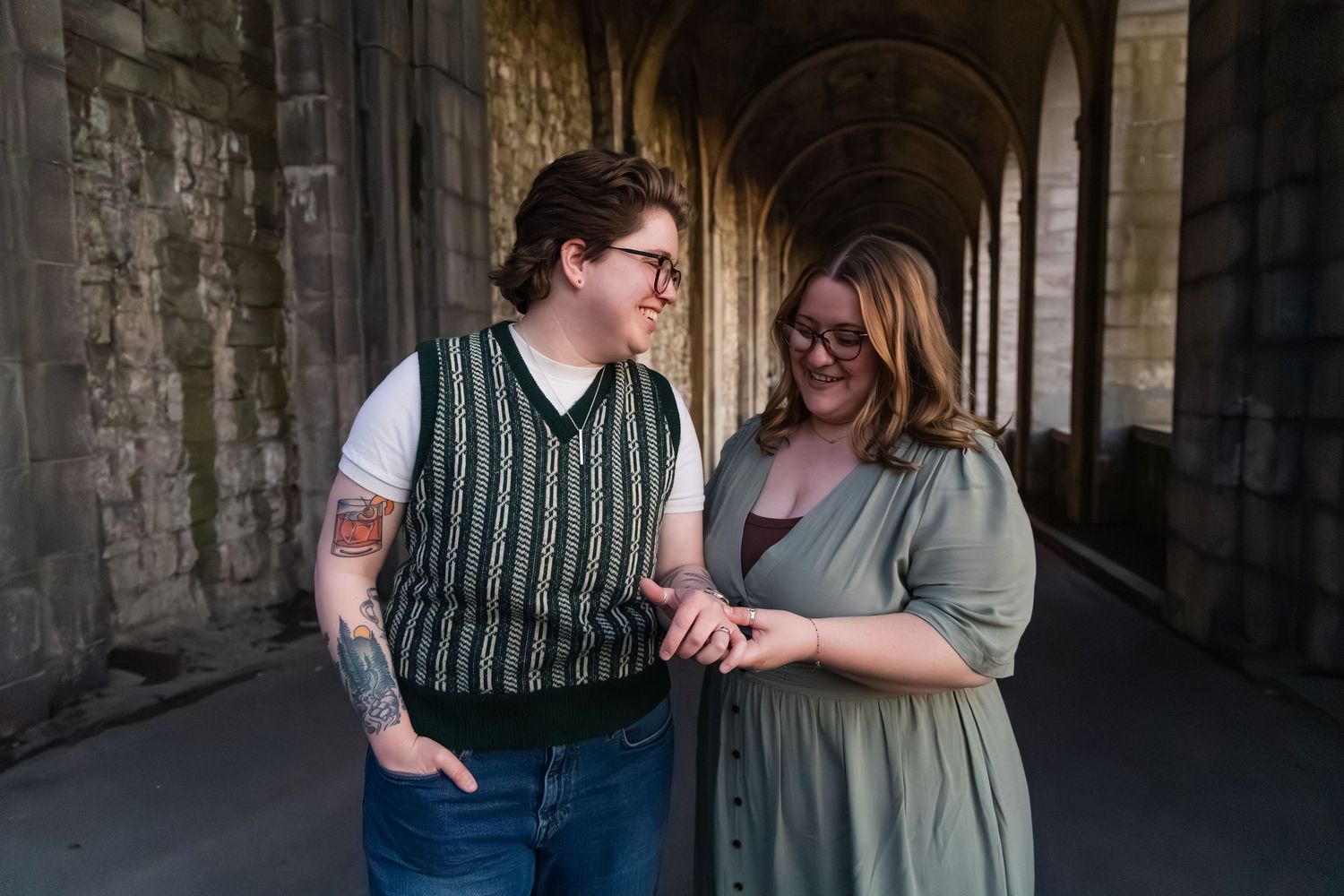 Couple smiling at each other in a stone archway corridor, one wearing a green vest and the other a sage dress.