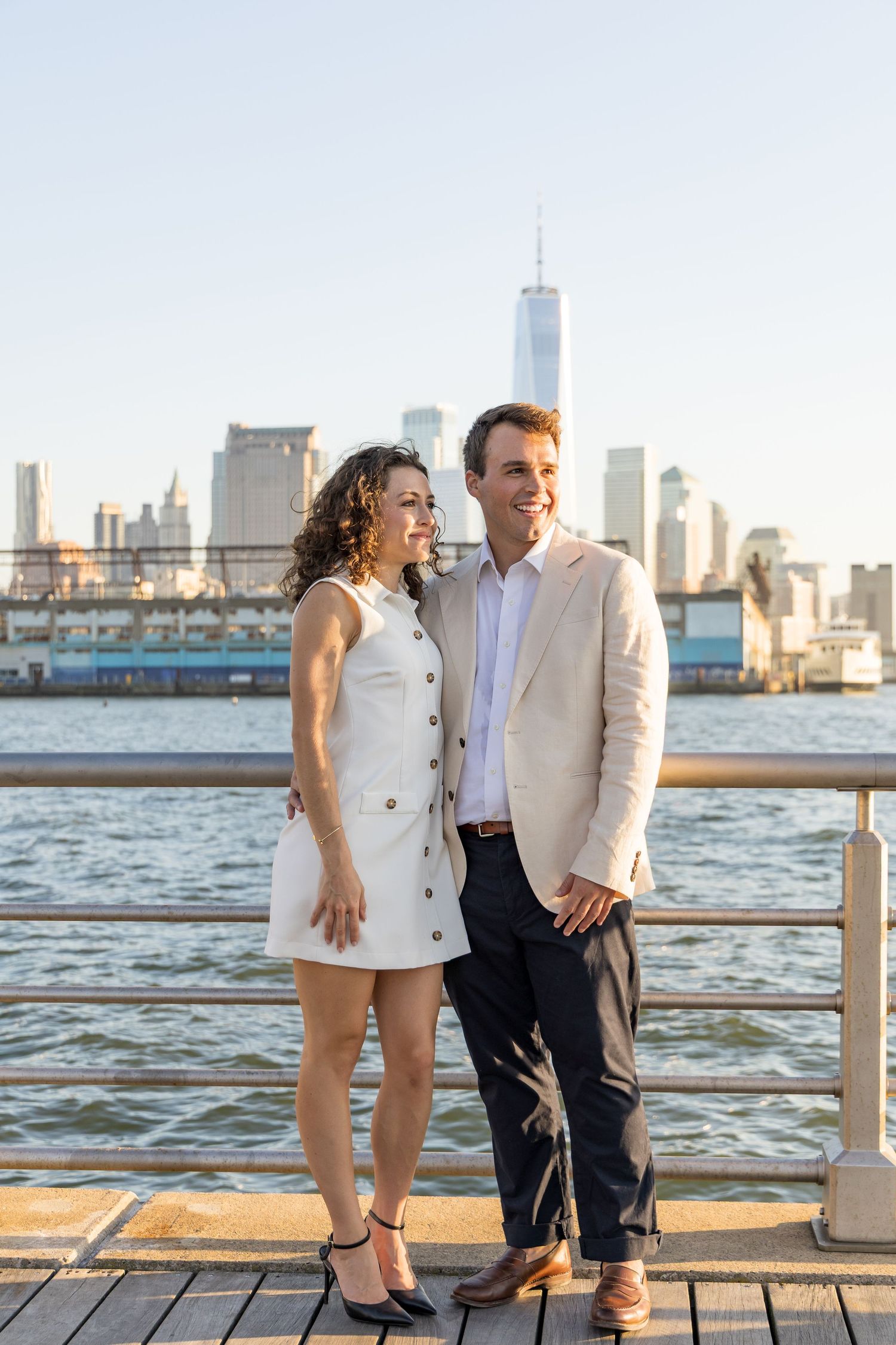 Couple posing on a waterfront pier with the New York City skyline and One World Trade Center in the background.