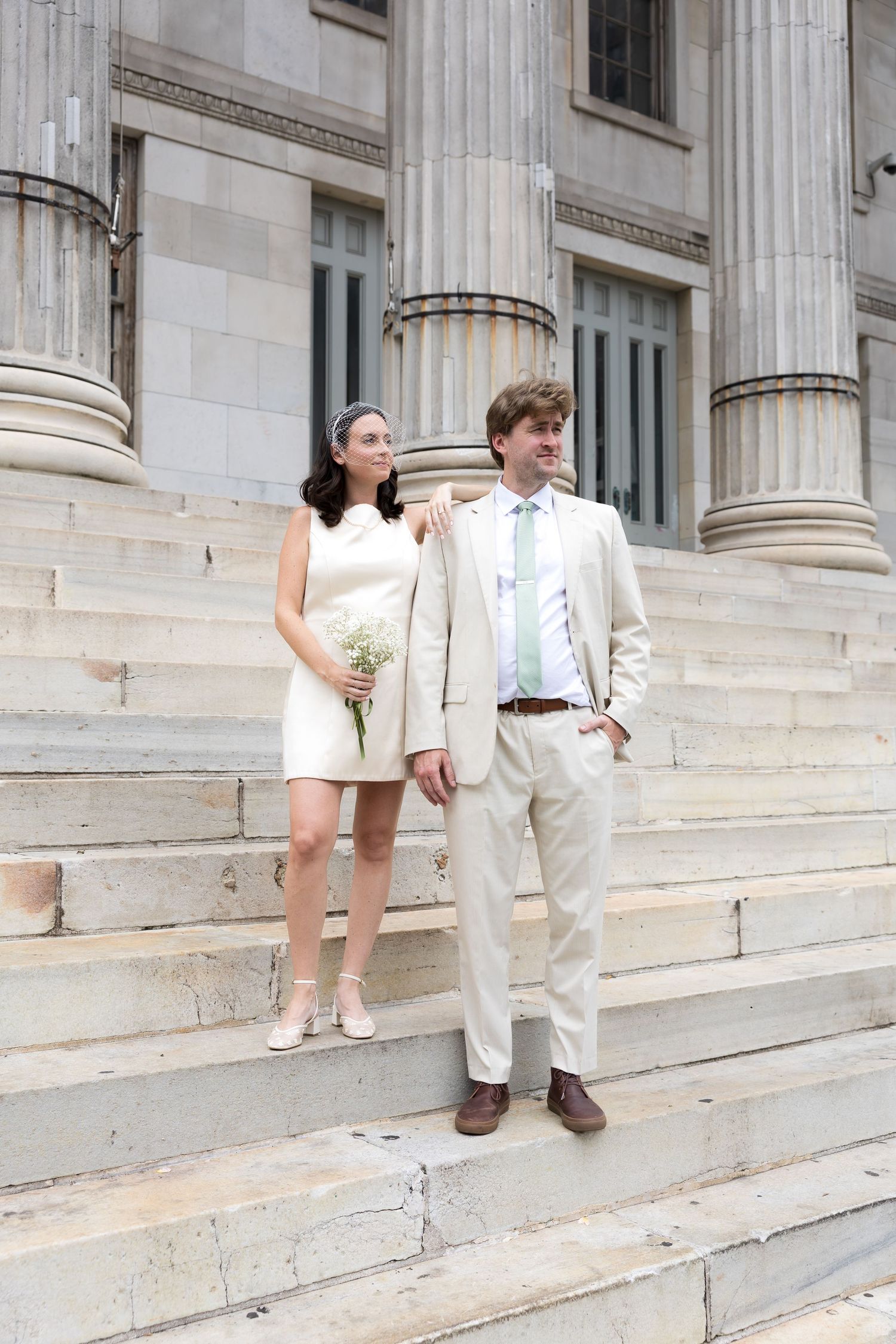 Bride and groom pose on stone steps of a grand building with columns, she holds white flowers.