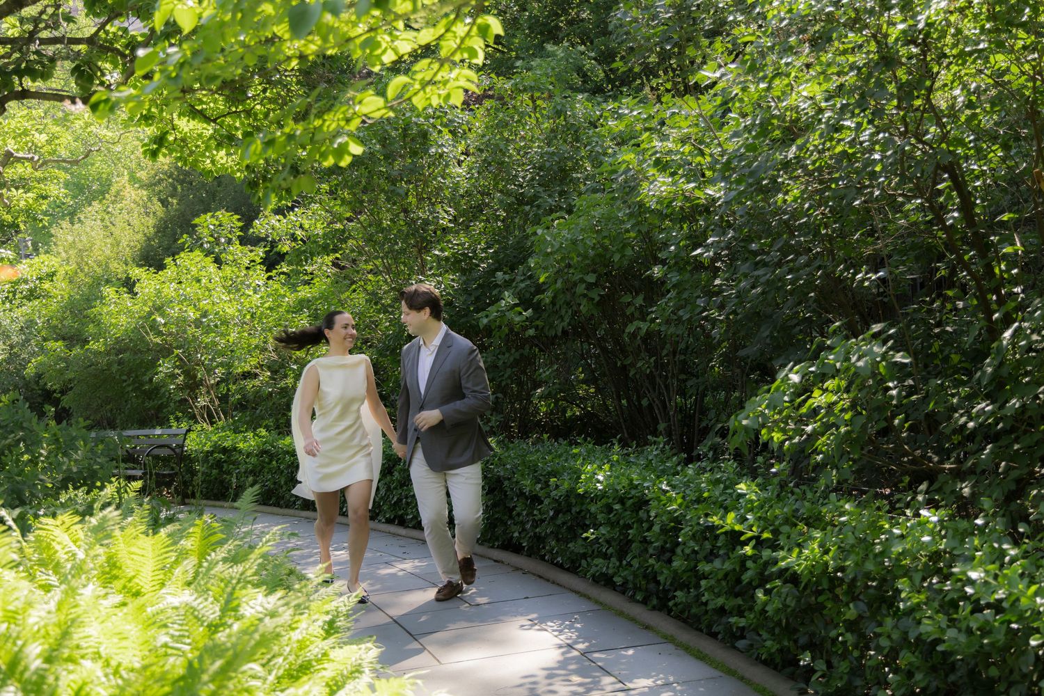 Couple walking hand in hand on a scenic garden path surrounded by lush green trees and shrubs.