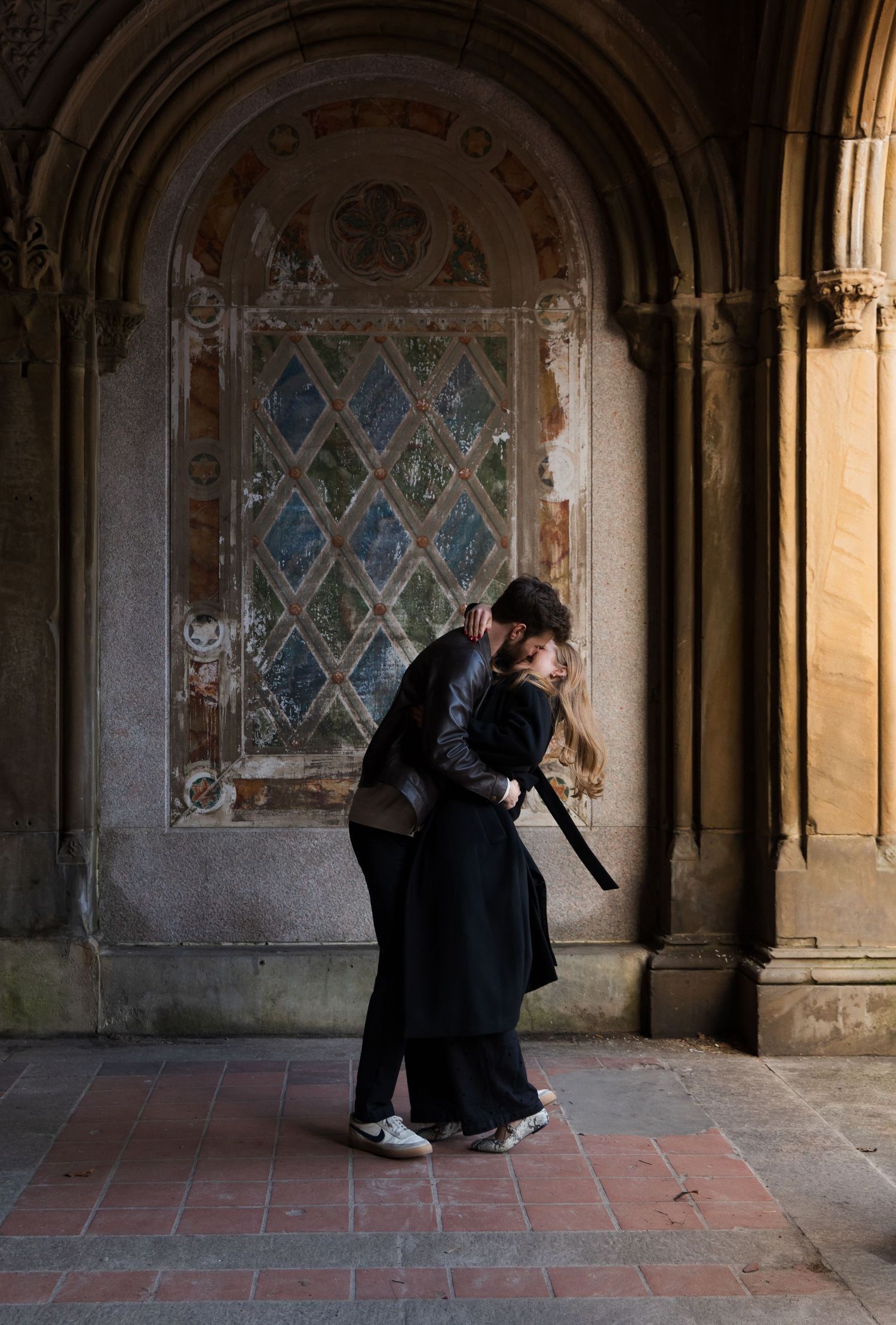 Couple embracing and kissing in an ornate stone archway with decorative frescoes in a historic building.