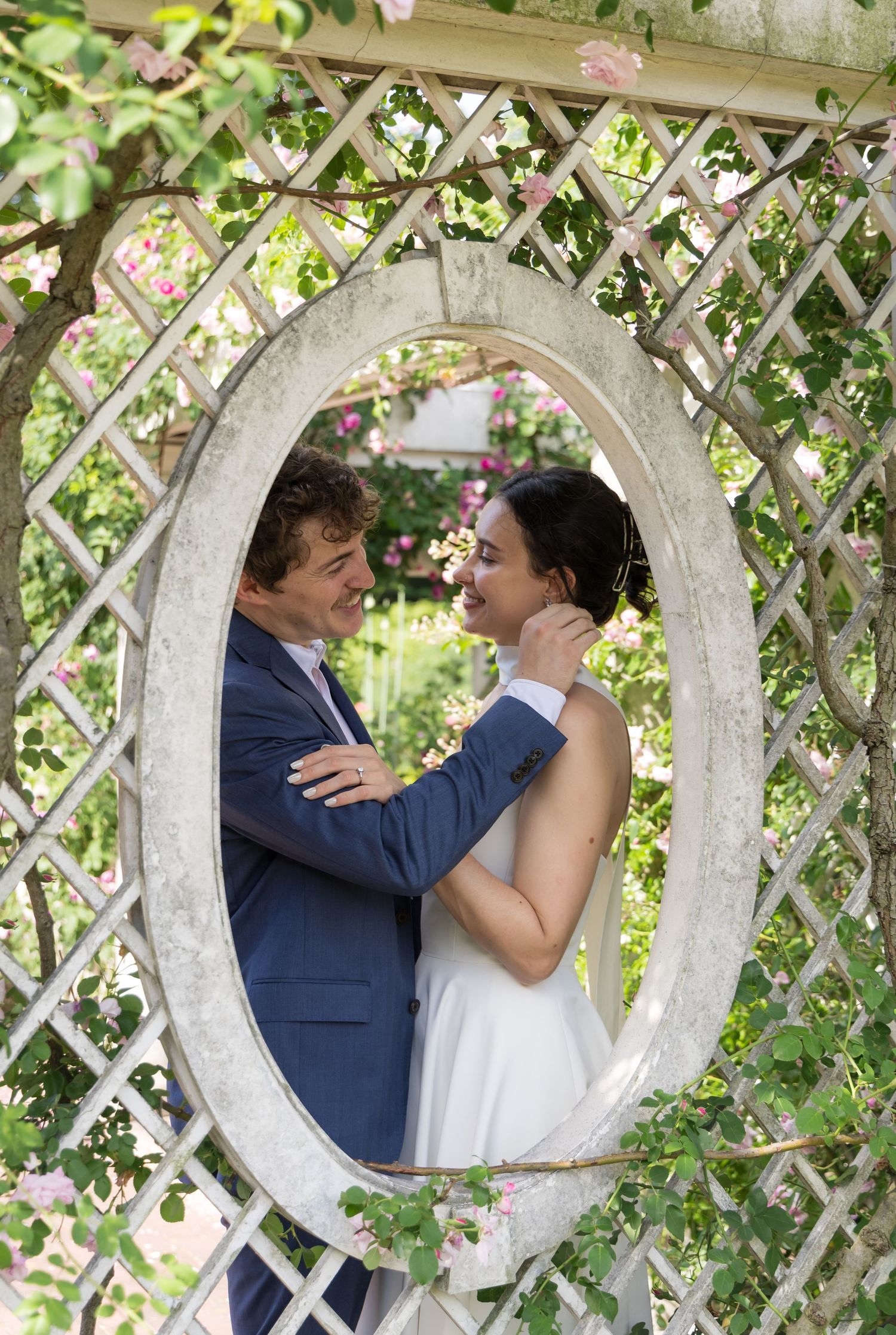 Bride and groom share a romantic moment framed by an oval garden trellis surrounded by lush greenery and pink roses.