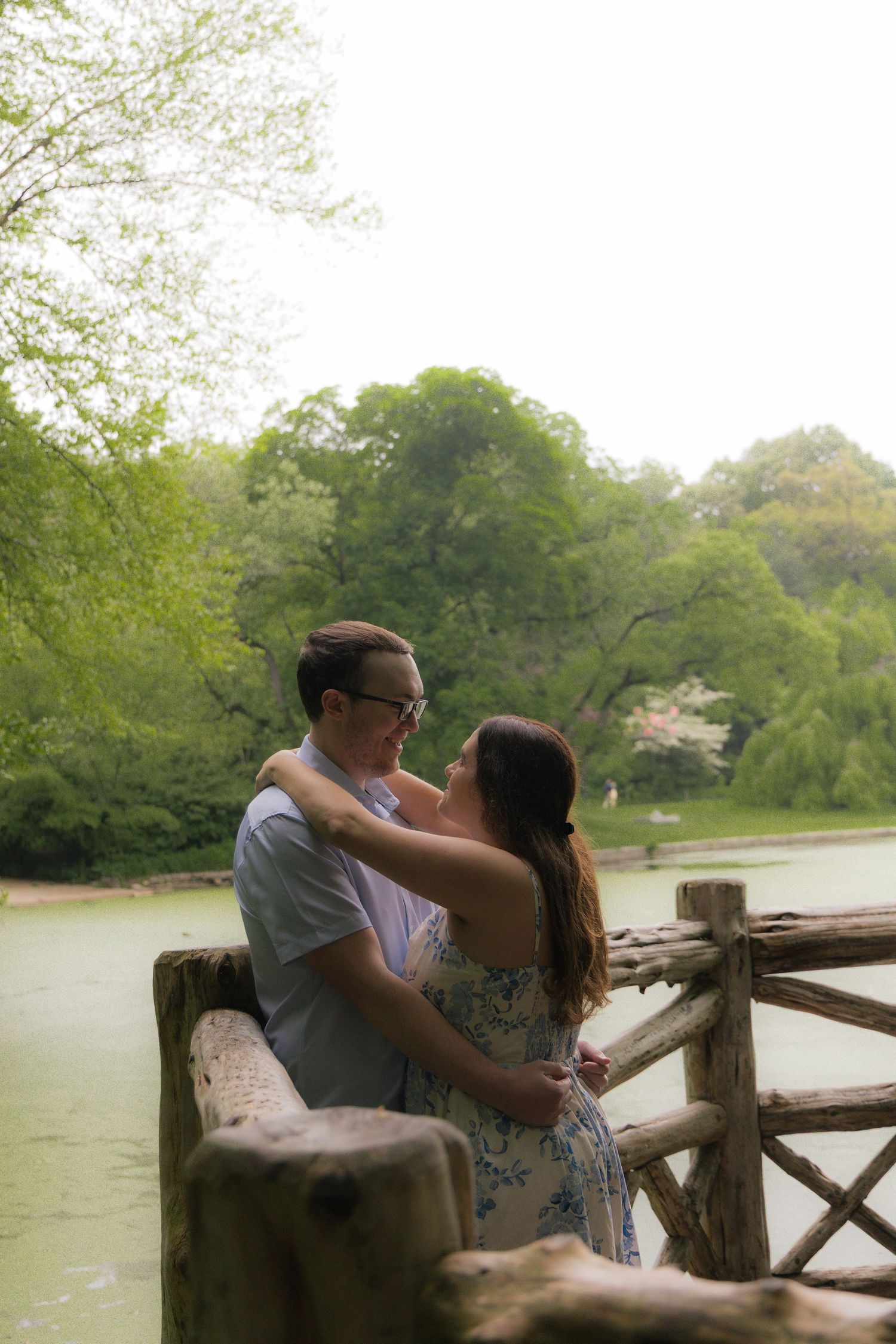 Couple embracing on a rustic wooden bridge overlooking a serene pond surrounded by lush green trees in a park.