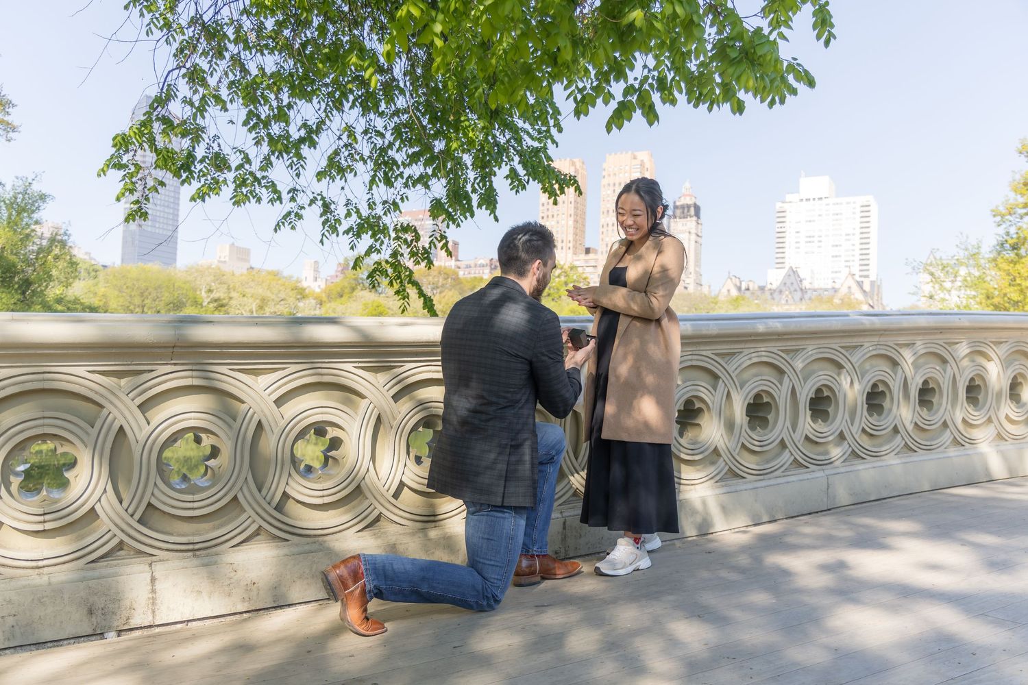 Man proposes on one knee to woman on Bow Bridge in Central Park NYC with city skyline in background.