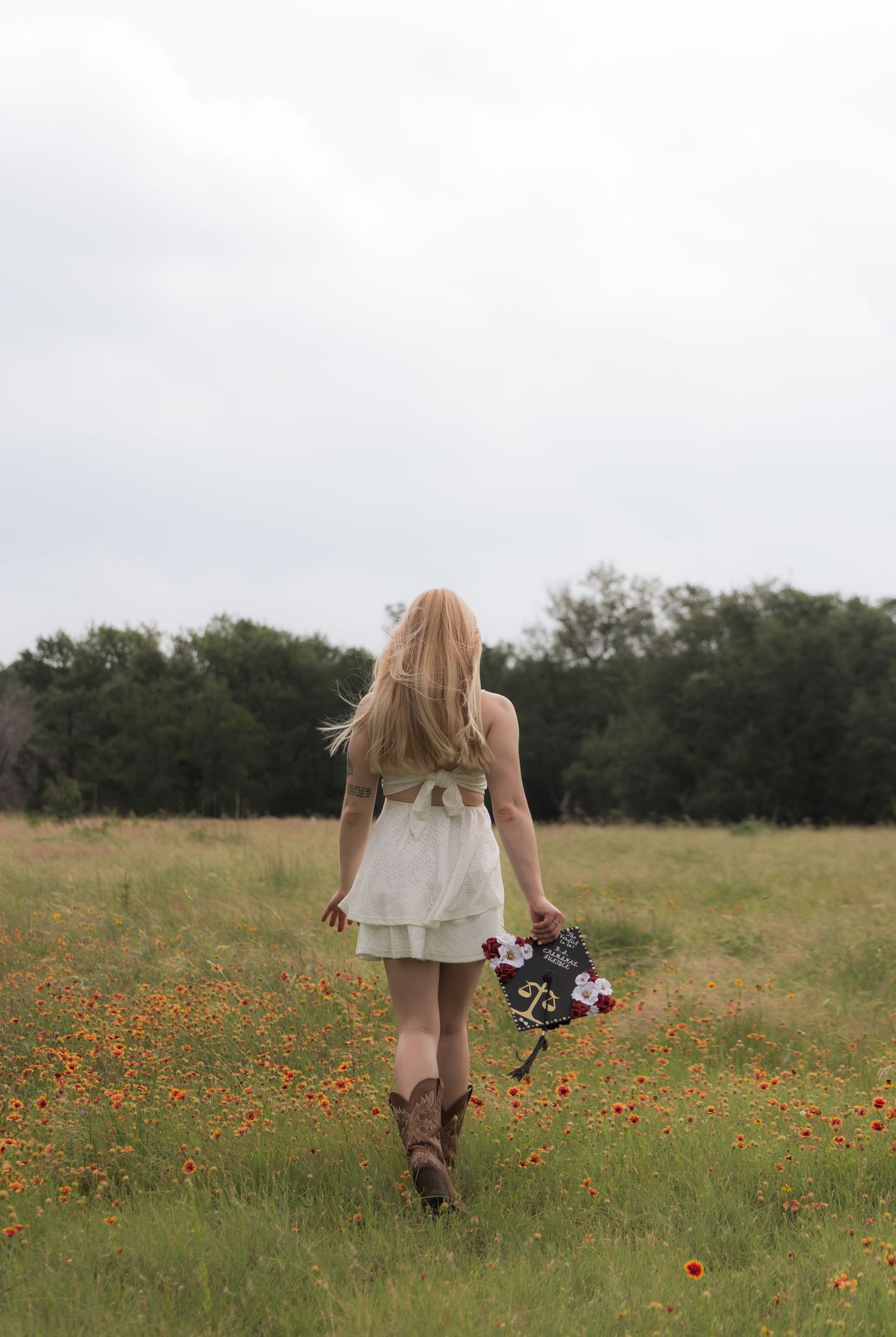 Rear view of blonde woman in white dress and cowboy boots walking through orange wildflower field.