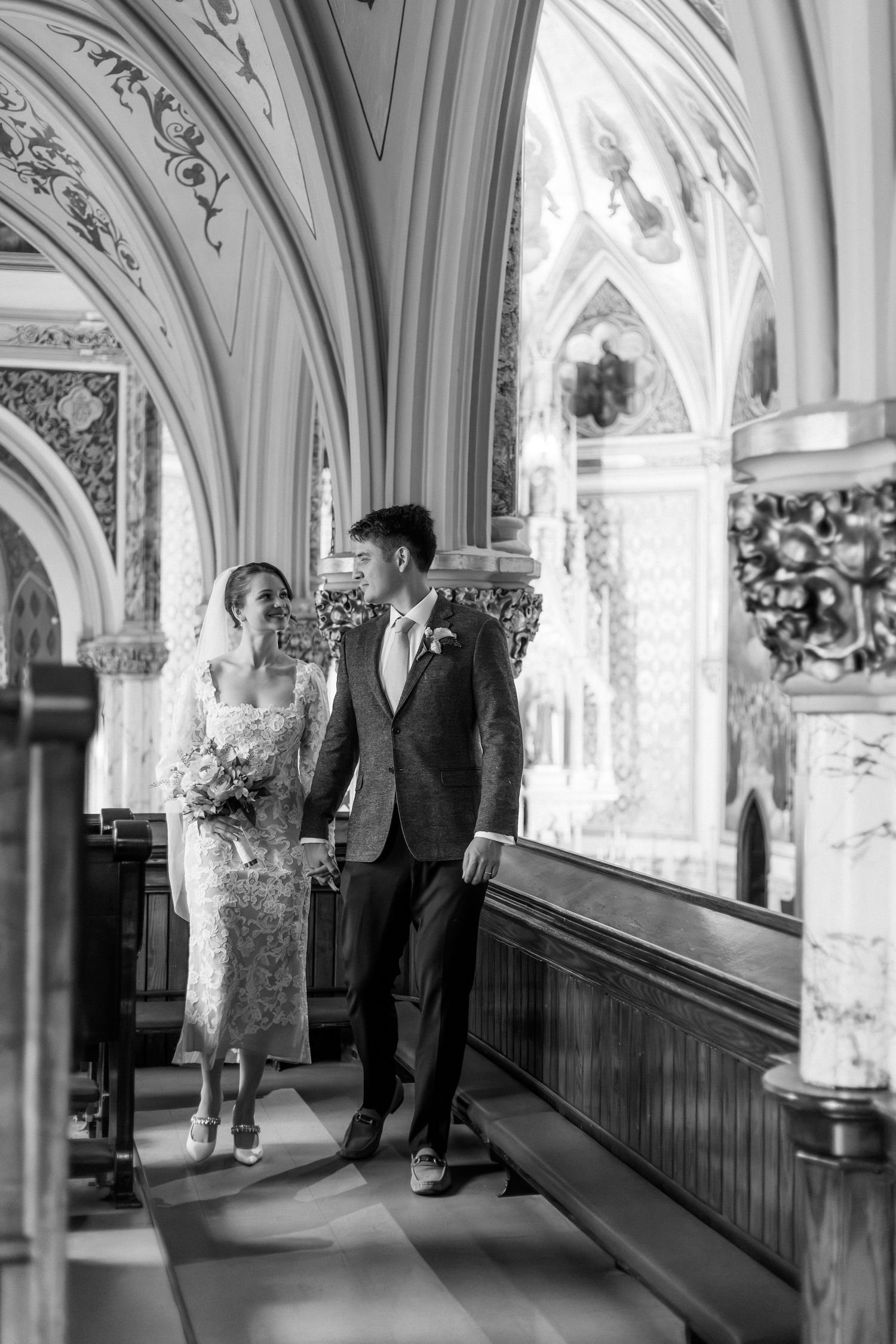 Black and white photo of a bride and groom walking hand in hand down a grand church aisle after their wedding ceremony.