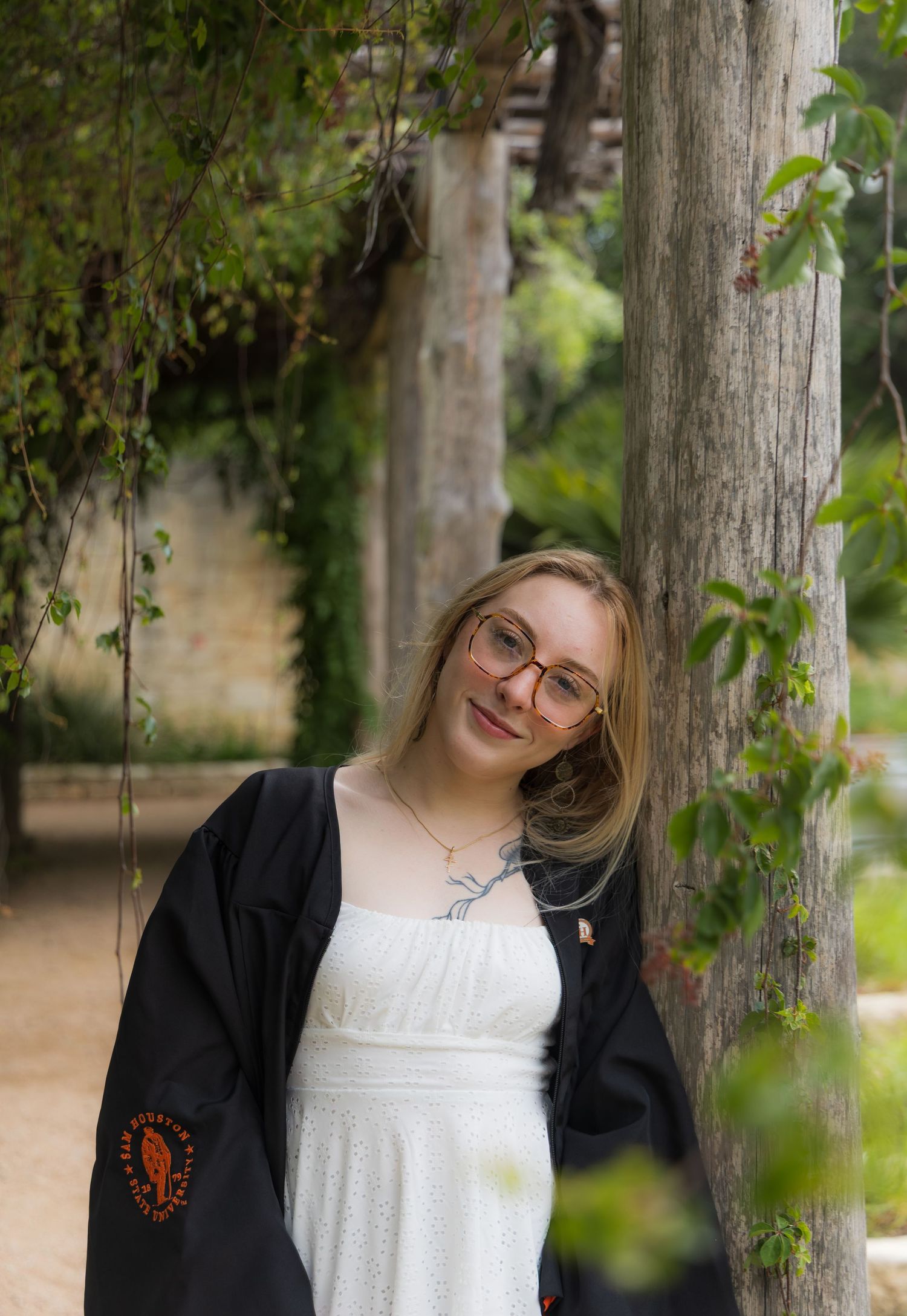 Smiling blonde woman in white dress and black graduation gown leaning against tree outdoors.