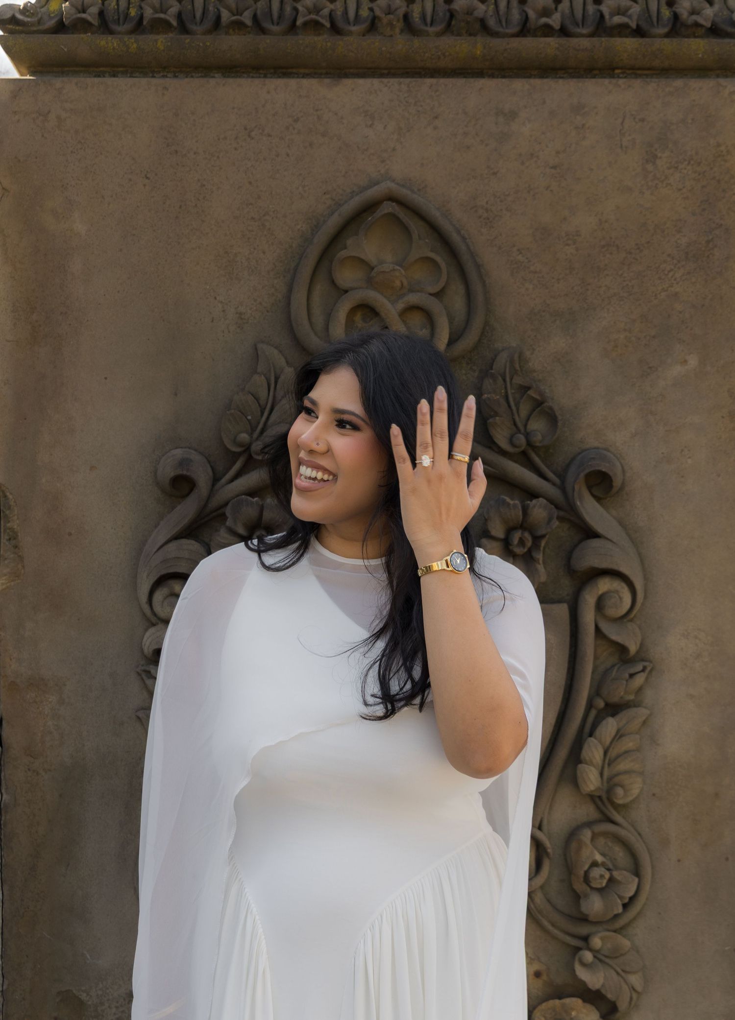 Smiling woman in white dress showing engagement ring, posed against ornate stone wall backdrop.