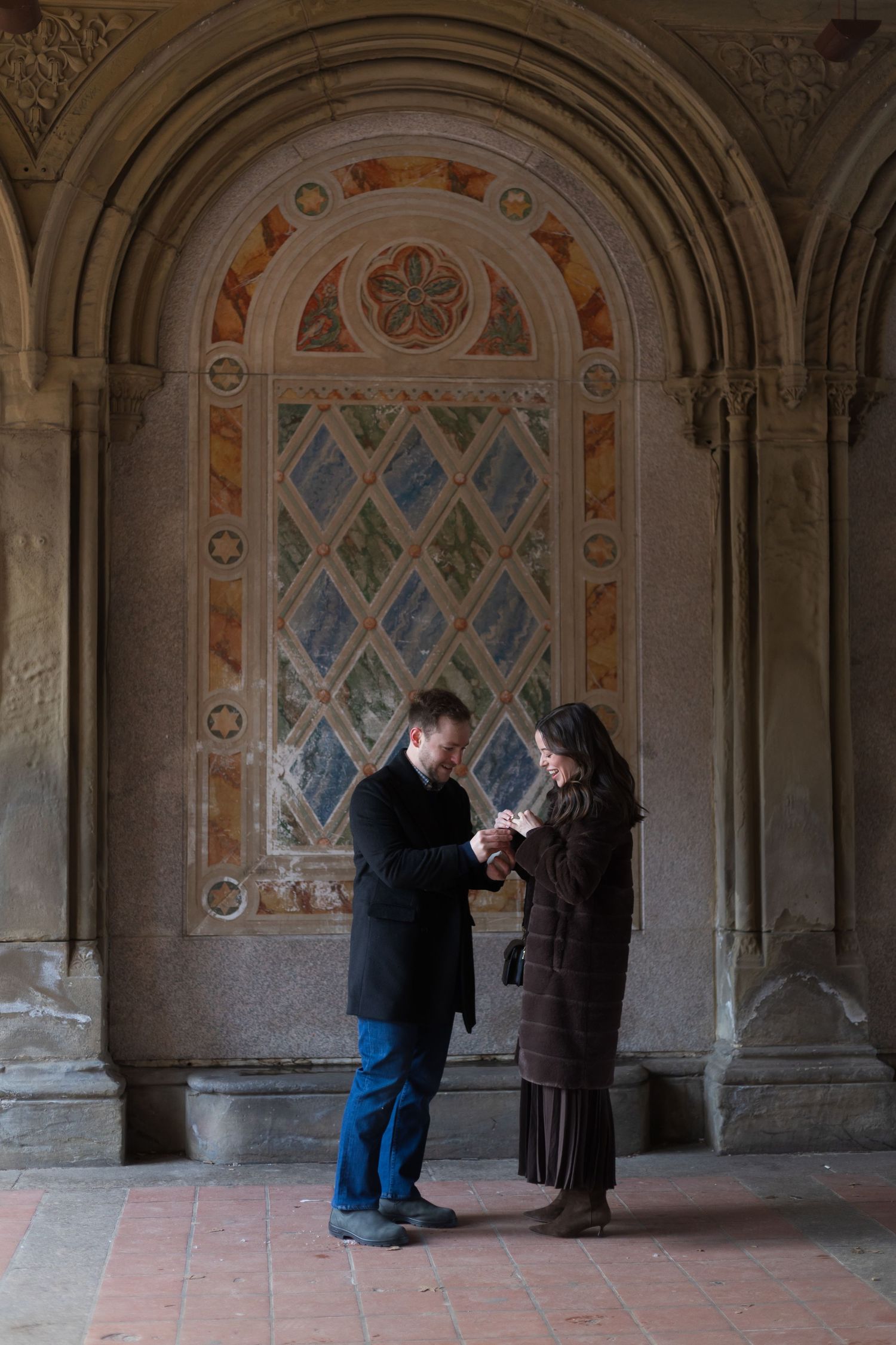 A man putting an engagement ring on his now-fiancee under Bethesda Terrace at a surprise proposal photoshoot in NYC.