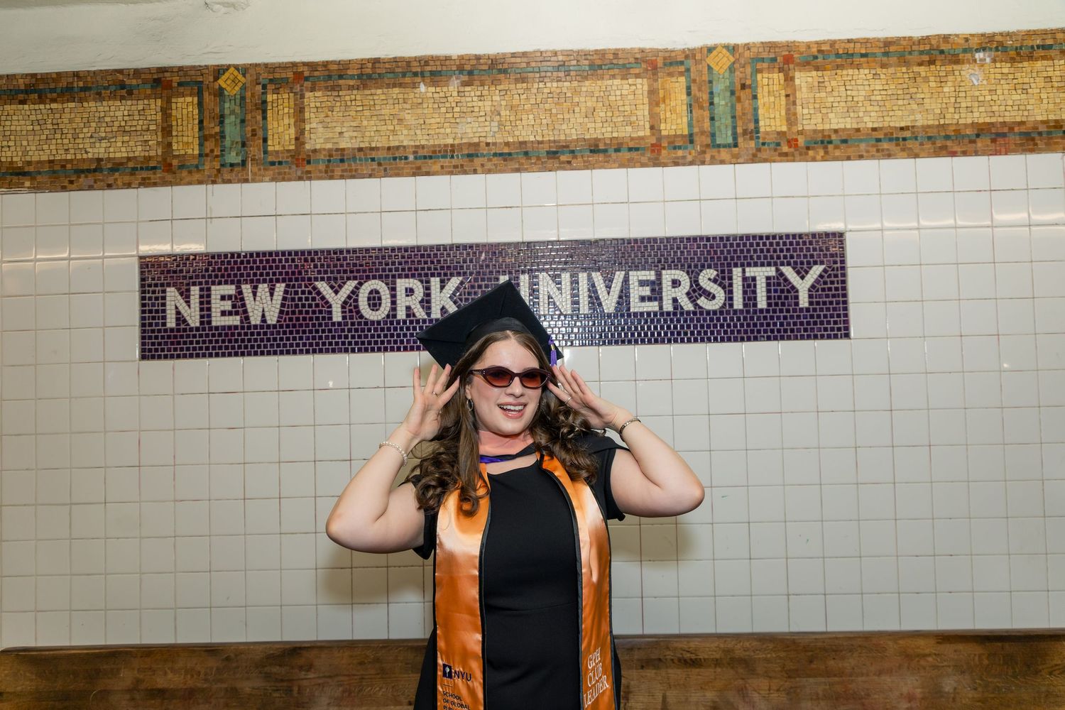 NYU graduate in cap and gown posing at New York University subway station tile sign in NYC.