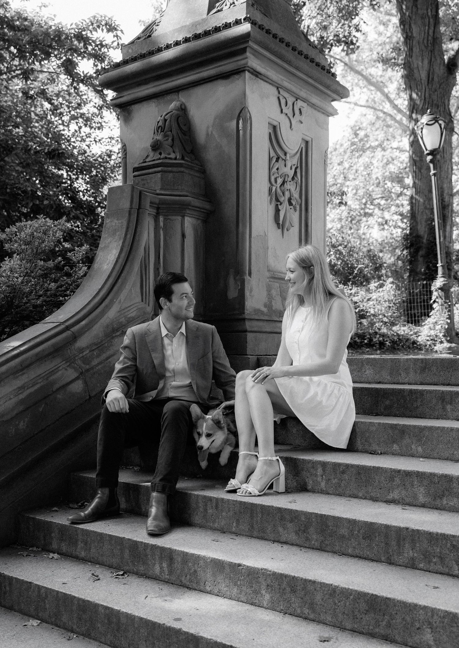 Couple sitting on stone steps with a corgi dog beside a decorative monument in a park, black and white photo.