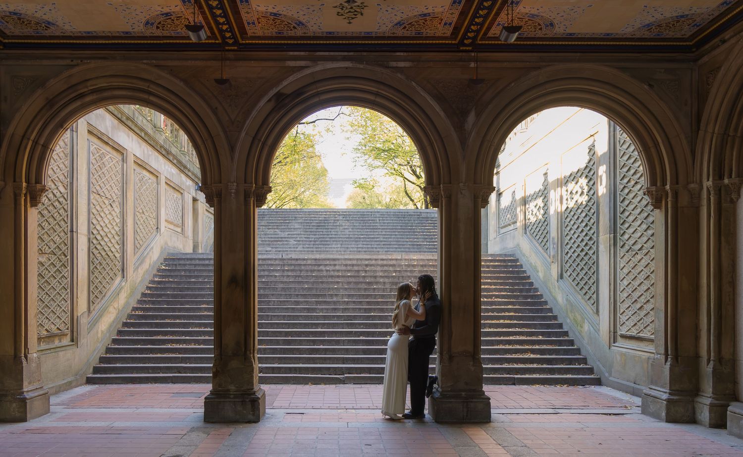 Couple embracing under Bethesda Terrace arches in Central Park, NYC, with grand staircases in background.