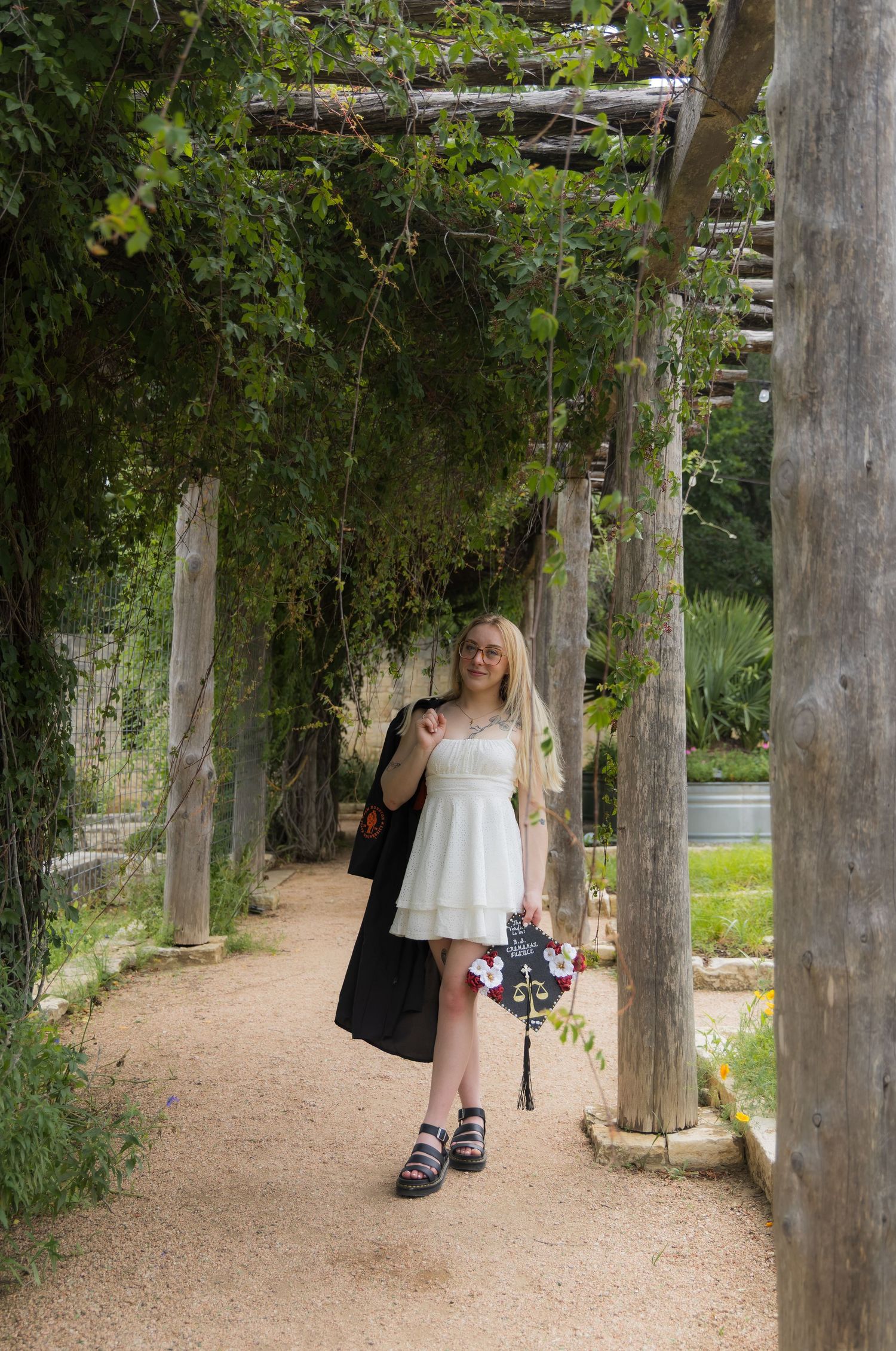 Blonde woman in white dress carrying black graduation gown under lush green pergola pathway.