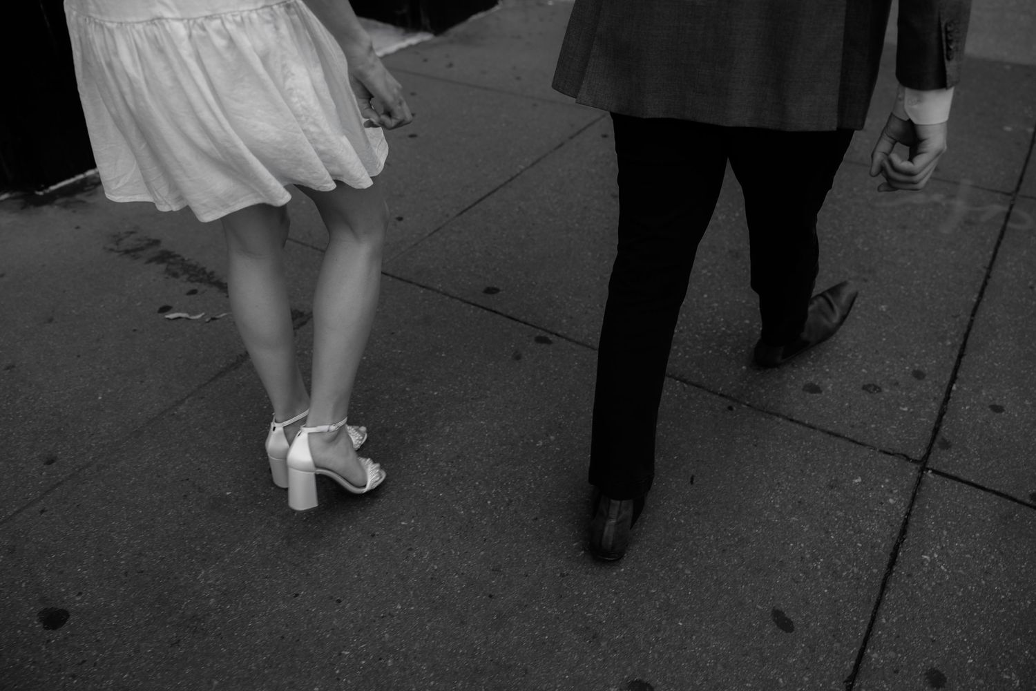 Black and white photo of a couple walking on a sidewalk, woman in white dress and heels, man in dark suit pants.
