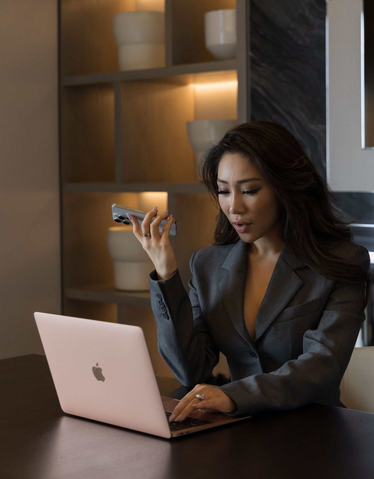 Professional in a gray blazer working on a MacBook laptop at a desk in moody lighting.