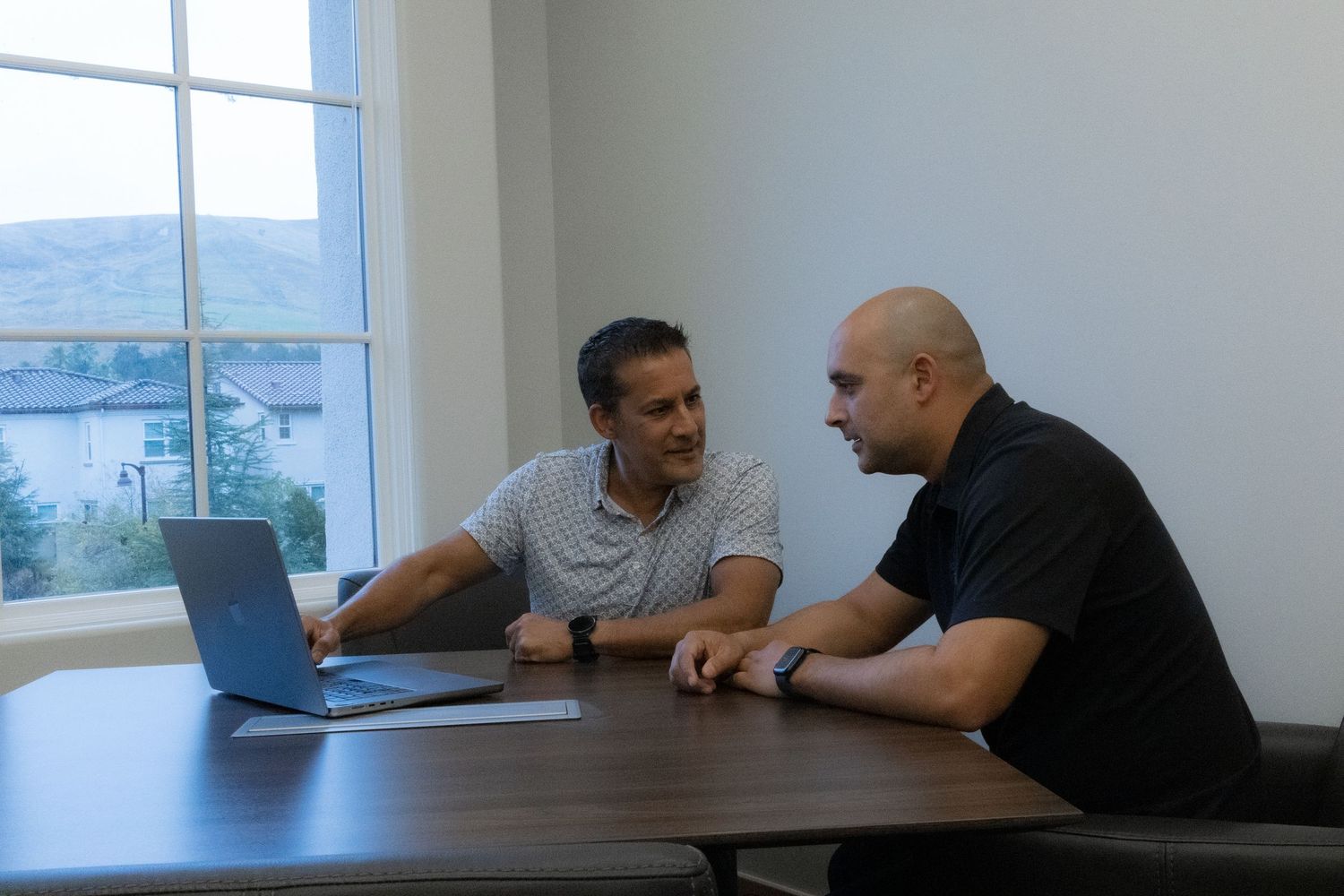Two people sit at a table in an office having a business discussion with natural light coming through windows.