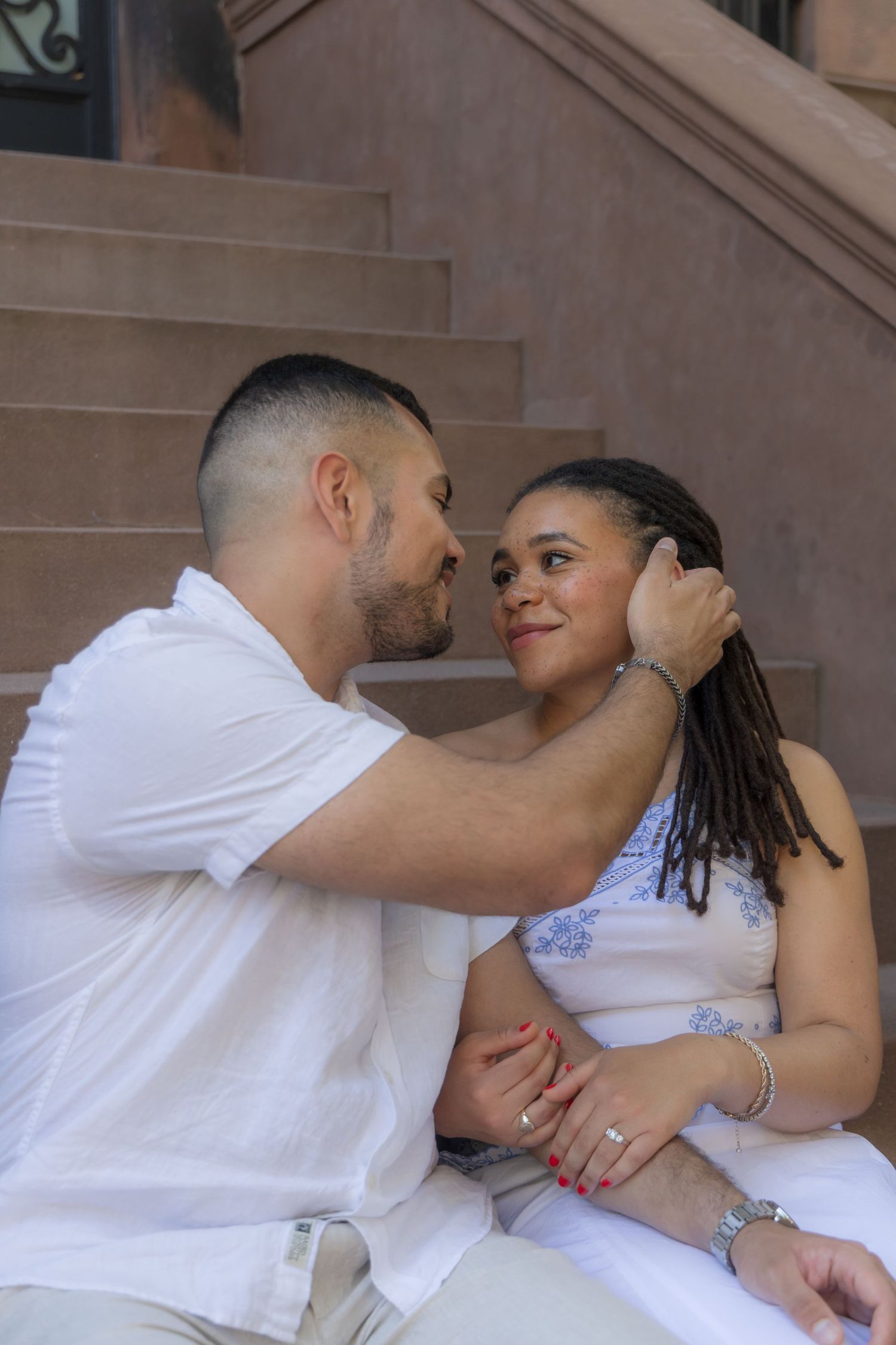 A couple sits on brownstone steps, man tenderly holds woman's face as they share an intimate moment.