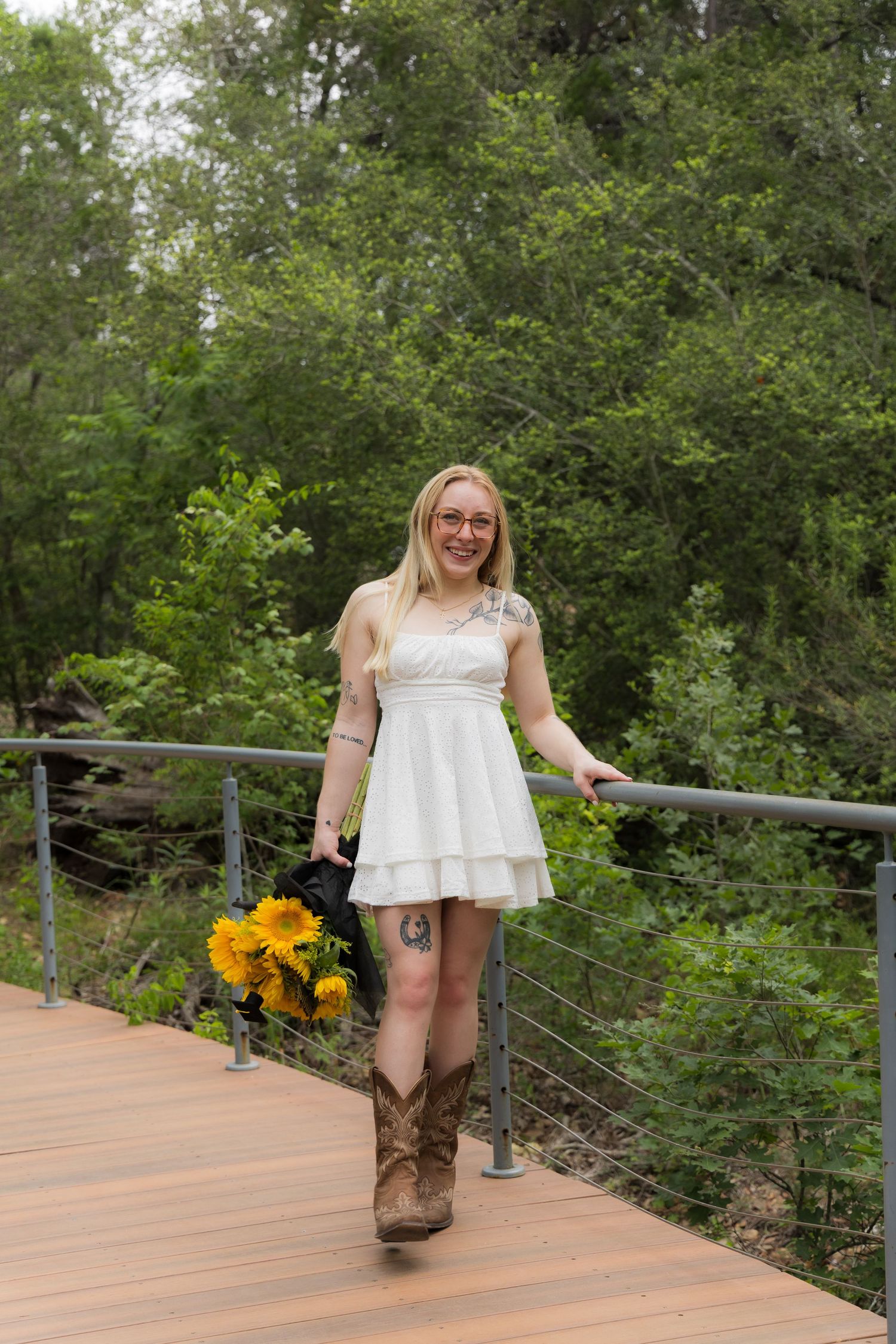 Smiling blonde woman in white dress and cowboy boots holding sunflowers on wooden bridge.
