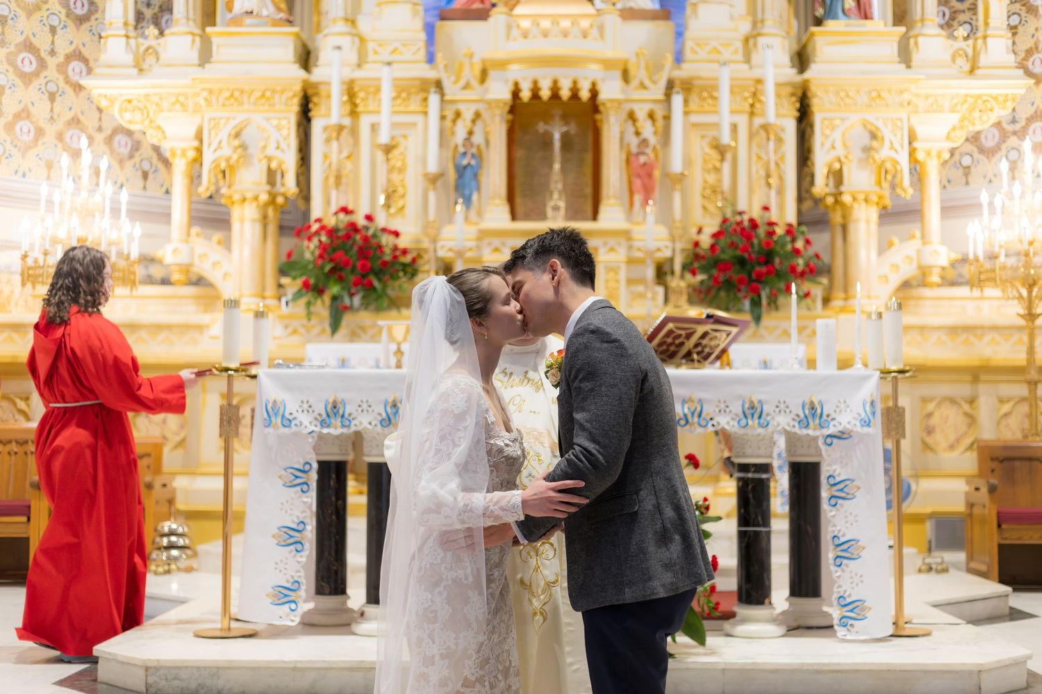 Bride and groom share a kiss at ornate church altar with red floral arrangements during wedding ceremony.