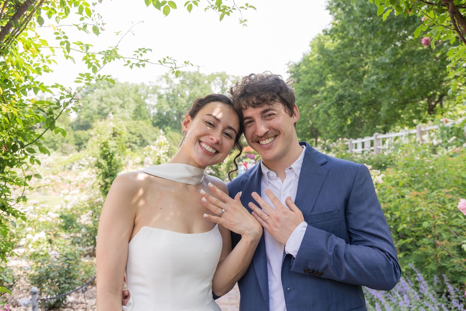 Newlywed couple smiling and showing wedding rings outdoors in a lush garden setting.
