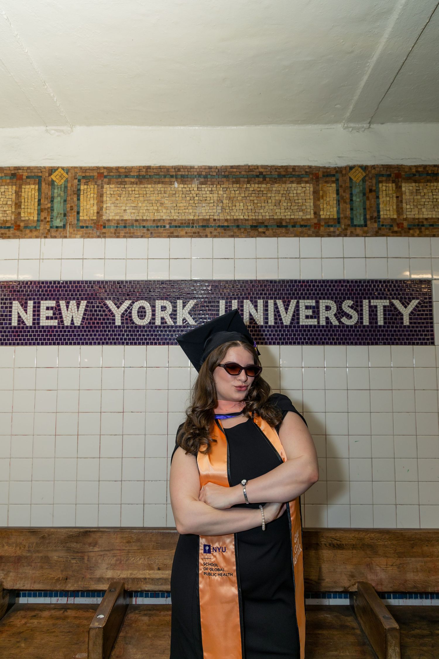 NYU graduate in cap and gown poses proudly at New York University subway station in NYC.
