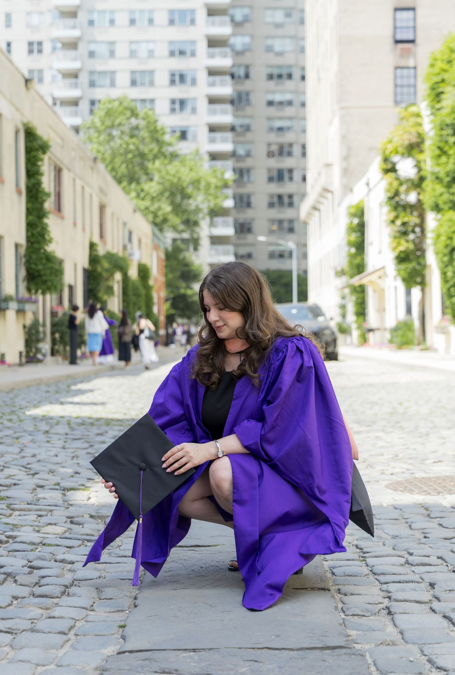 A woman in a purple graduation gown crouches on a cobblestone street holding her cap, with city buildings in the background.