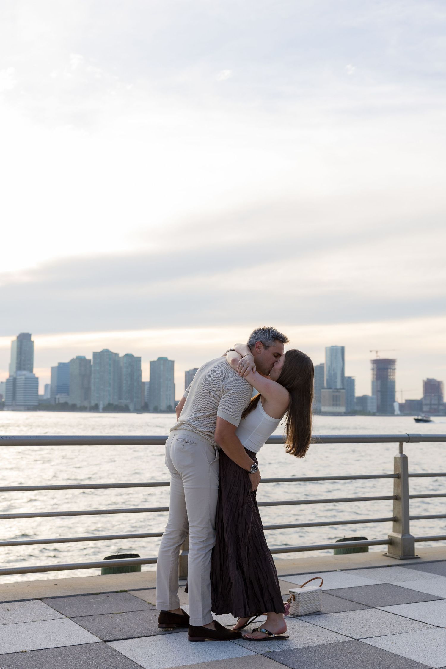Couple sharing a romantic kiss on a waterfront promenade with a city skyline and cloudy sky in the background.