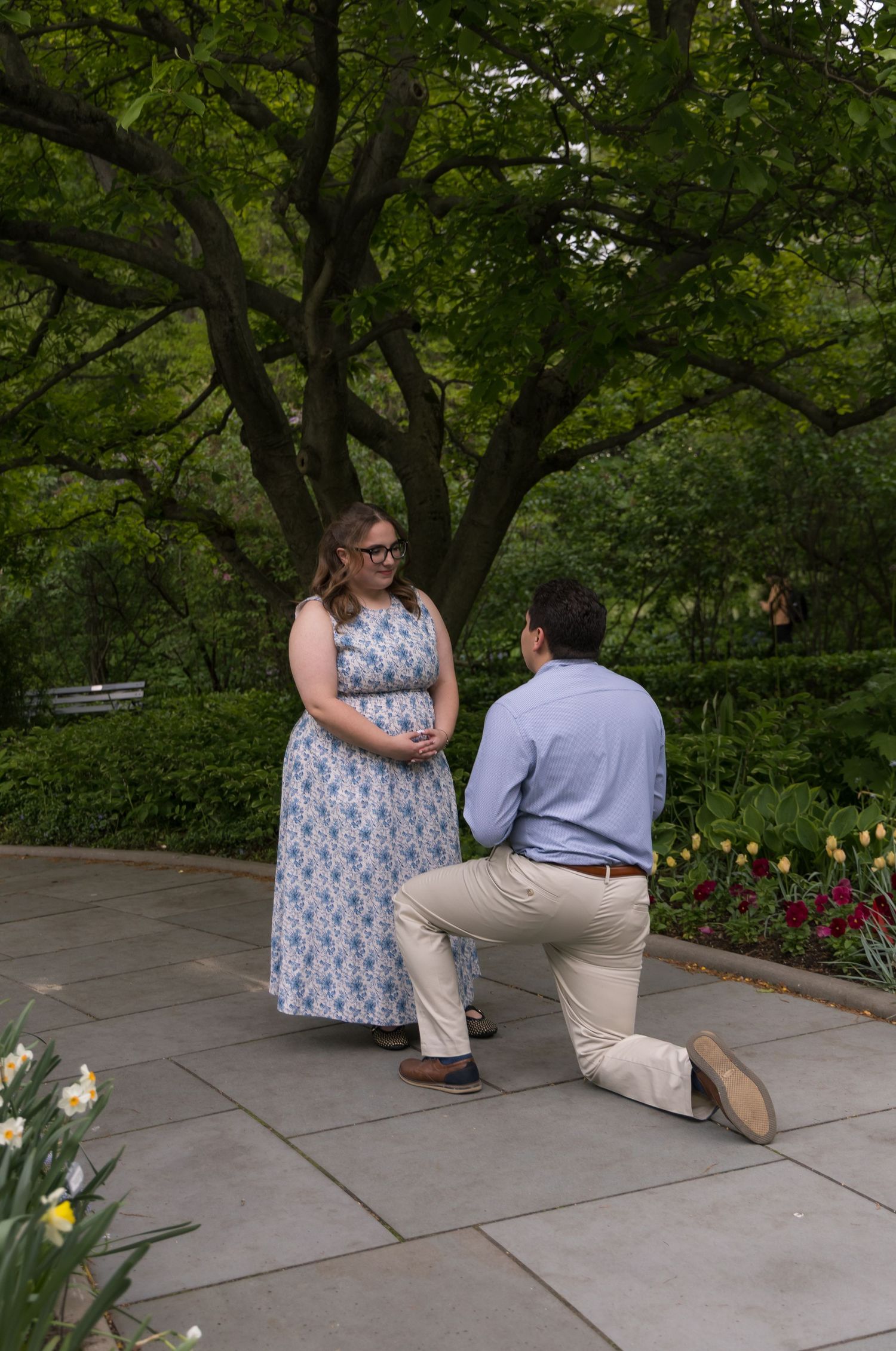 Man proposes on one knee to woman in floral dress in a lush garden park setting with tulips and trees.