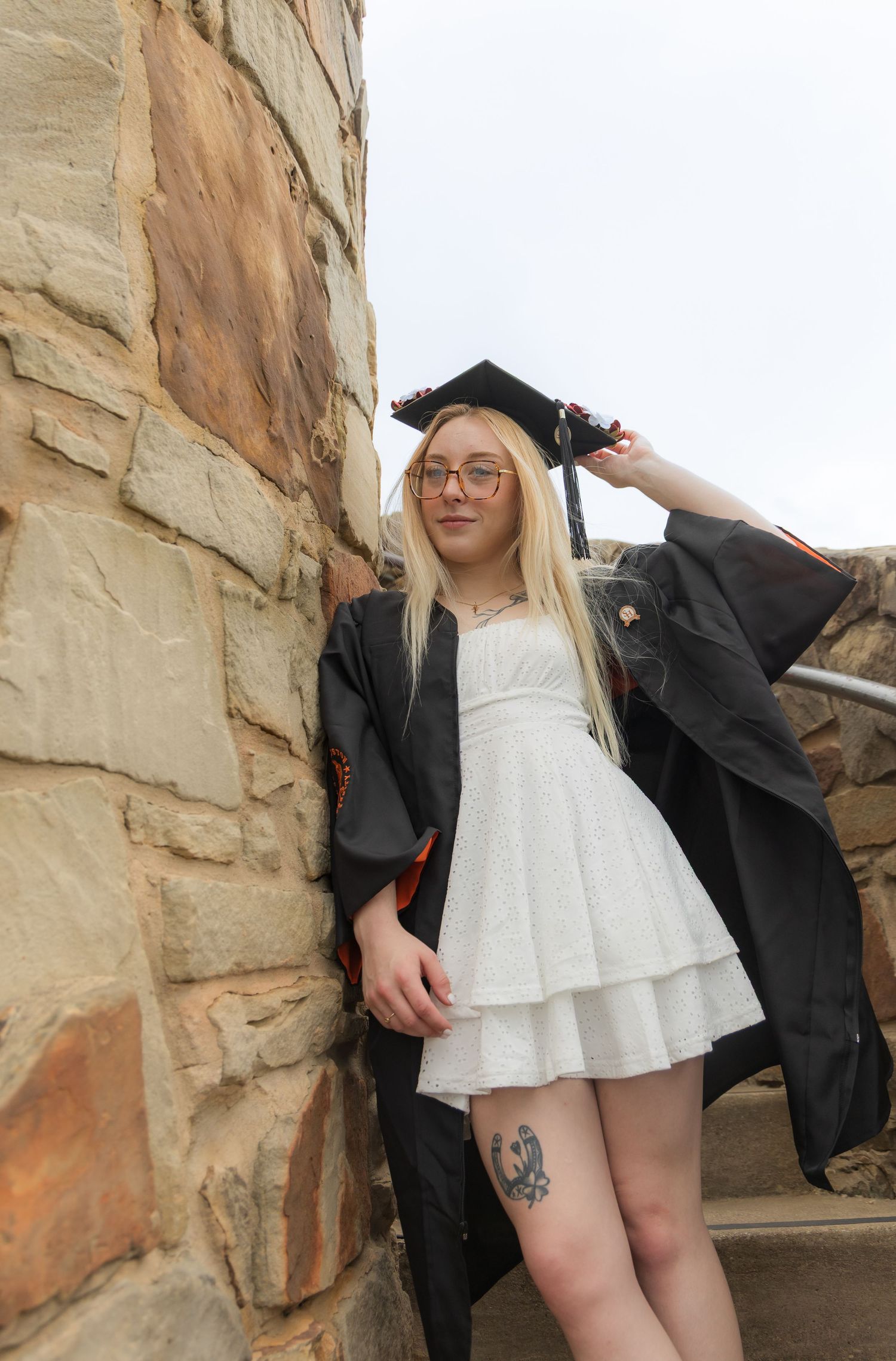 Blonde graduate in black cap and gown leaning against stone wall on outdoor staircase.