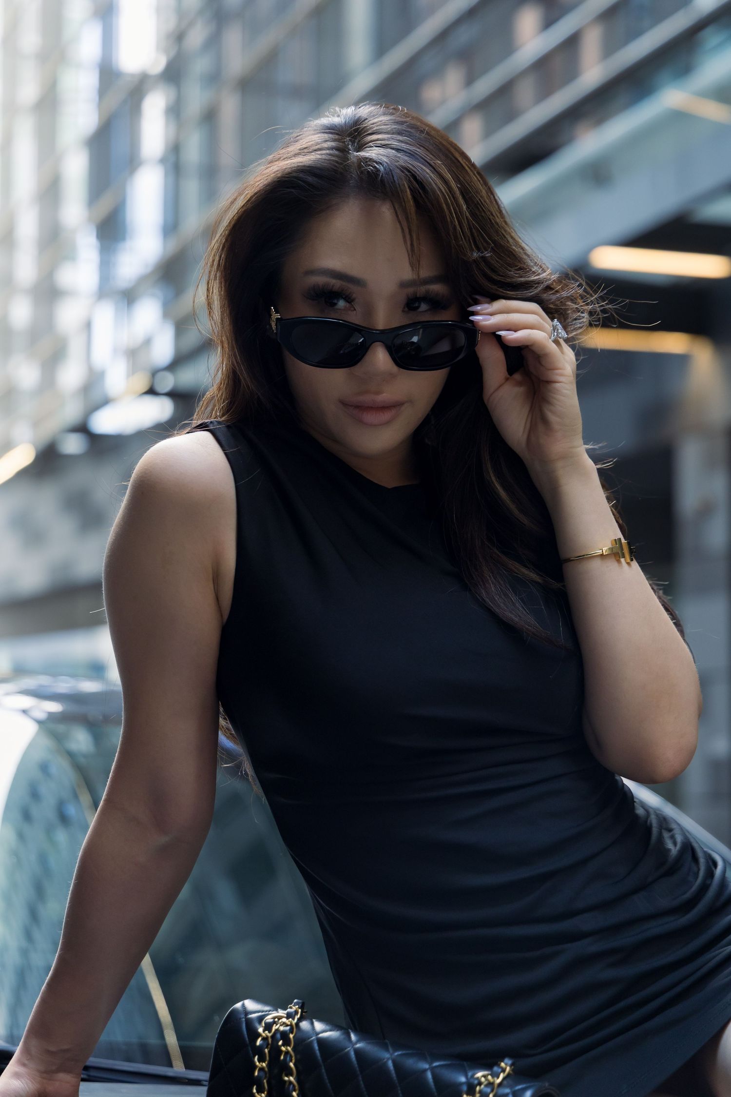 A woman in a black sleeveless top poses confidently with sunglasses in an urban setting.
