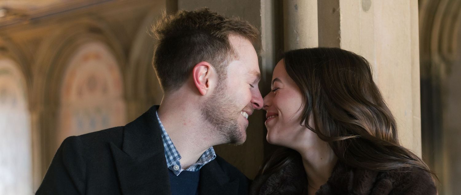 A close up  of a couple smiling nose-to-nose with the Bethesda Terrace archways in the background.