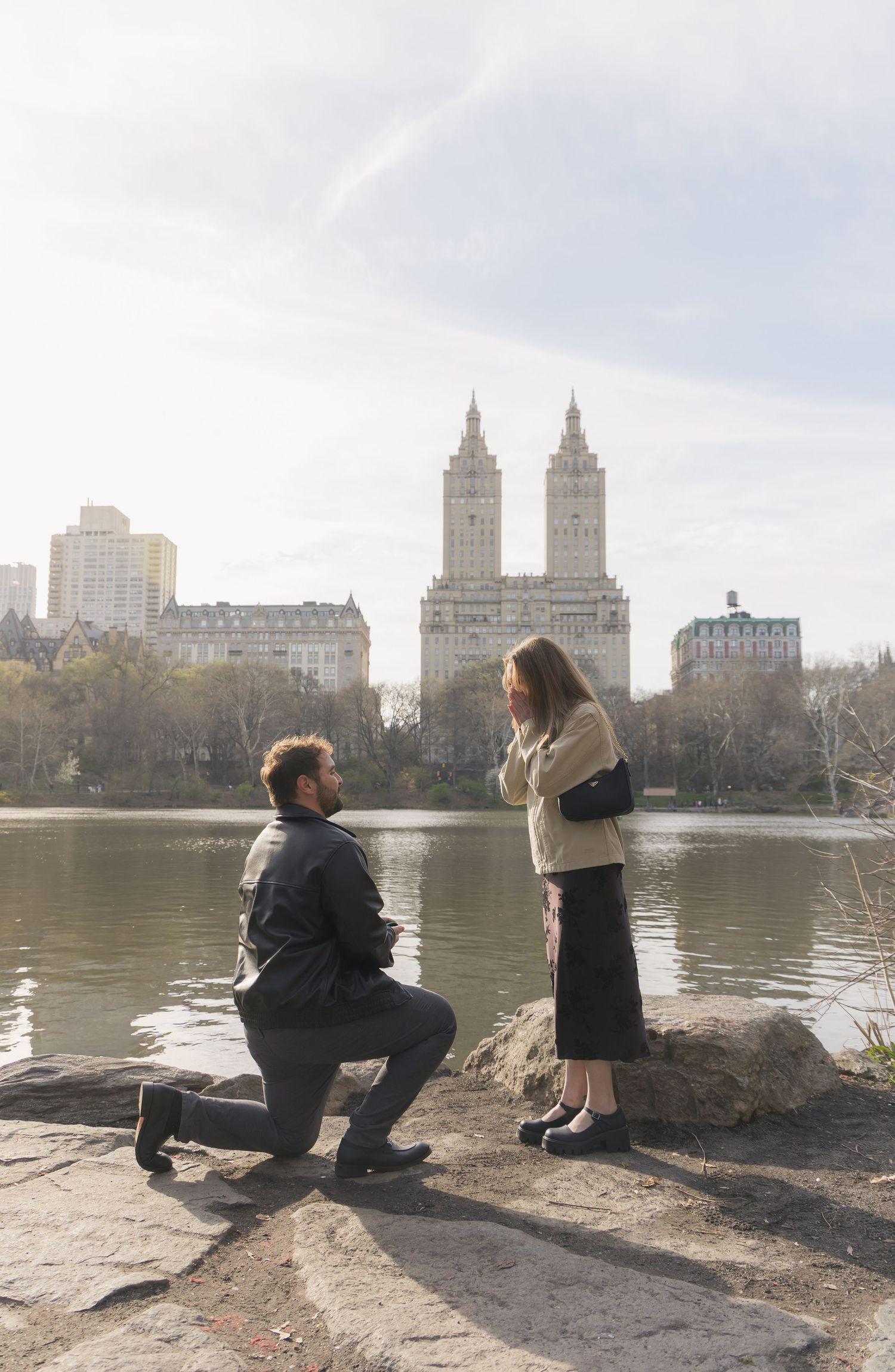 Man proposes on one knee to woman by Central Park lake with iconic San Remo building skyline in background.