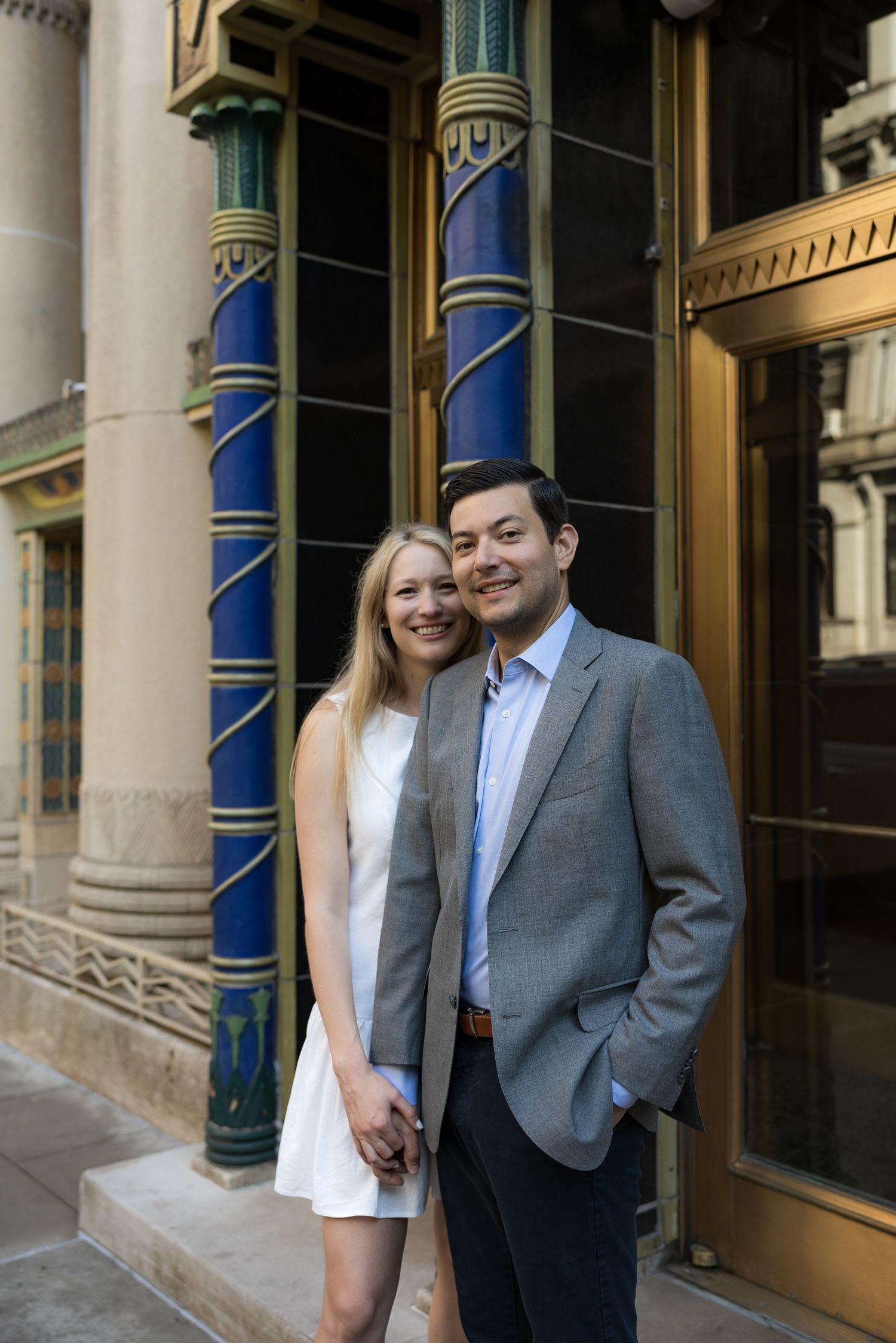 Couple posing together in front of ornate blue and gold architectural columns in an elegant urban setting.