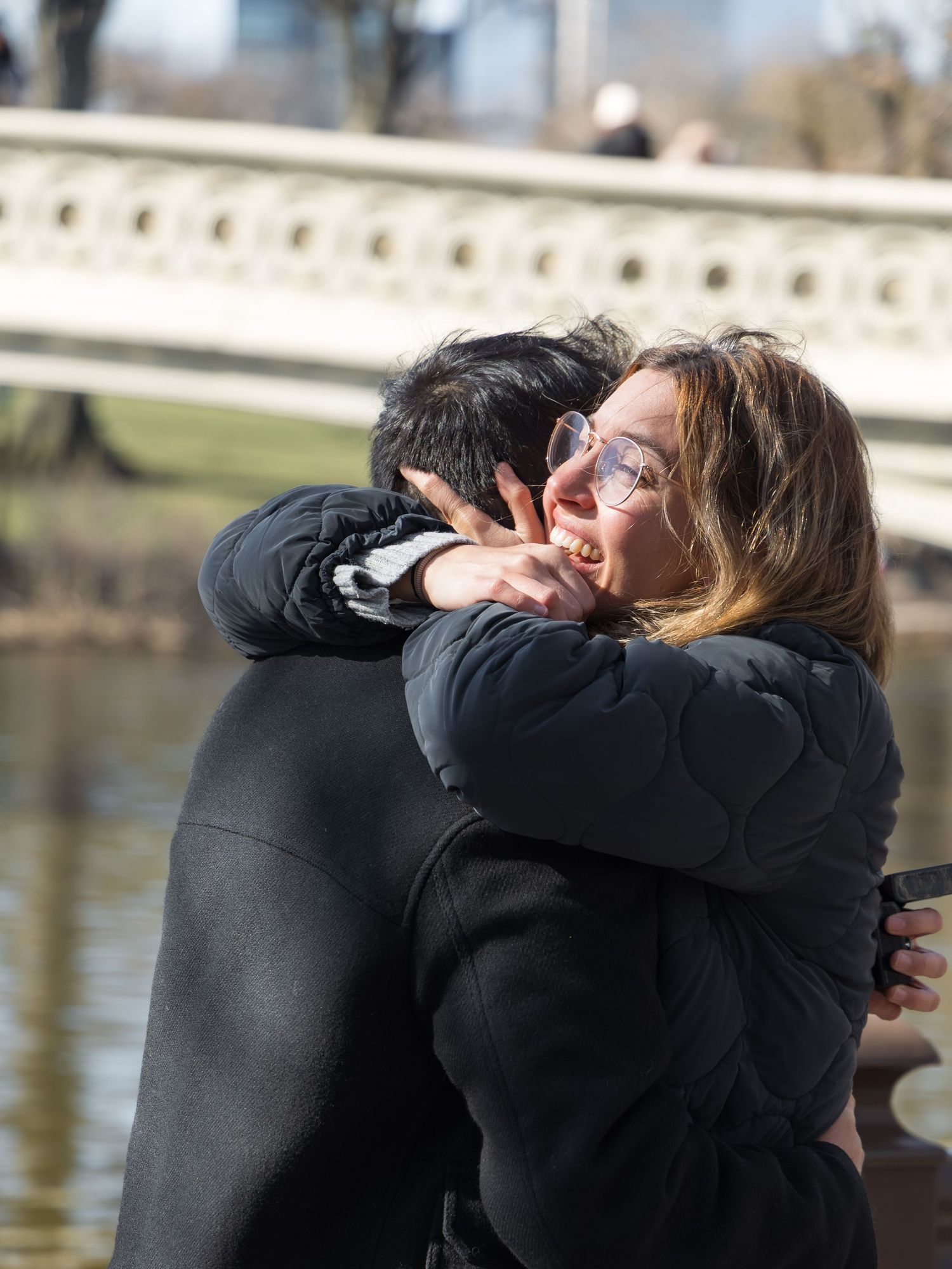 Emotional couple sharing a joyful embrace outdoors near a bridge, woman wearing glasses crying happy tears.