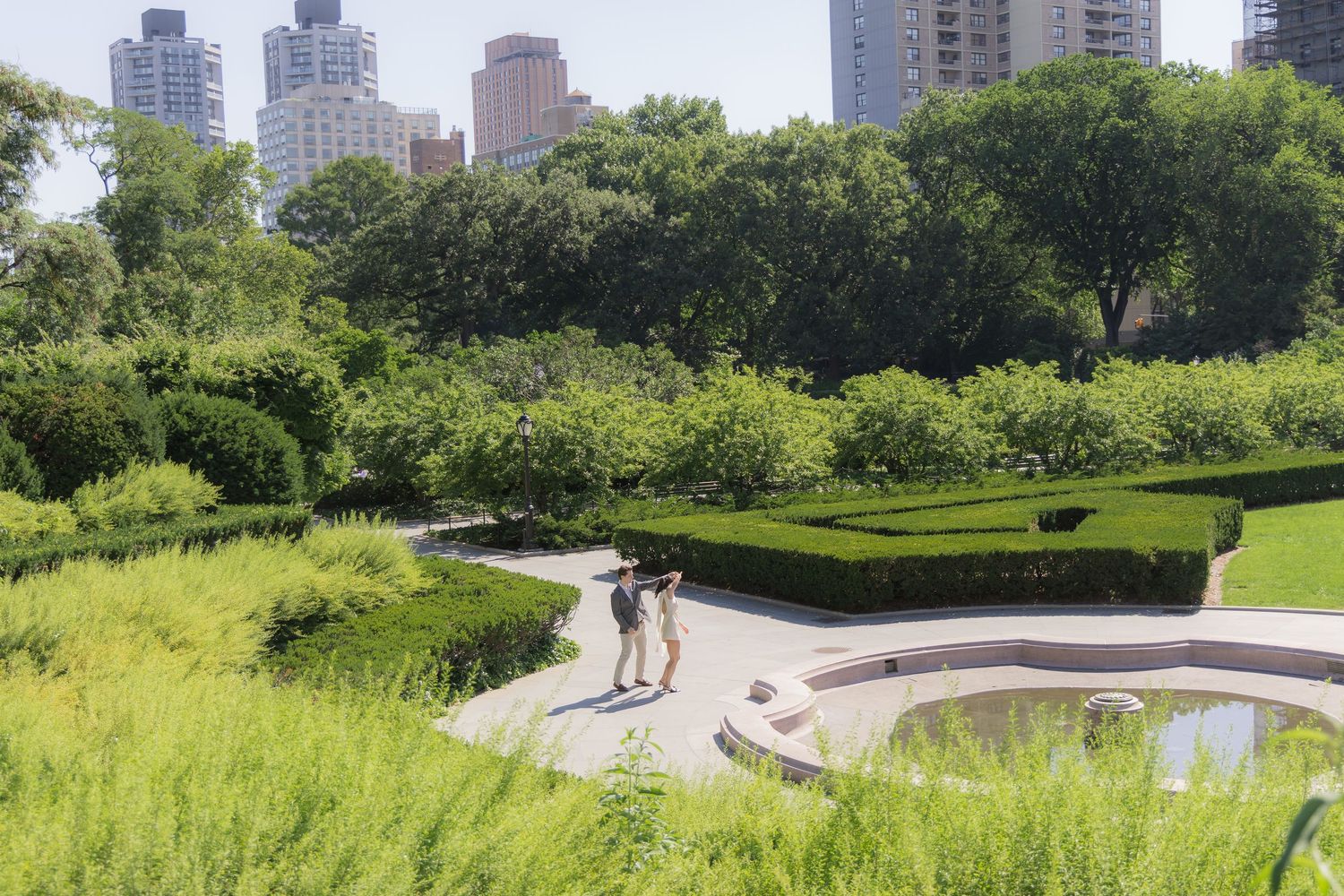 Two visitors stroll along a curved pathway in a lush urban garden with manicured hedges and city skyline beyond.