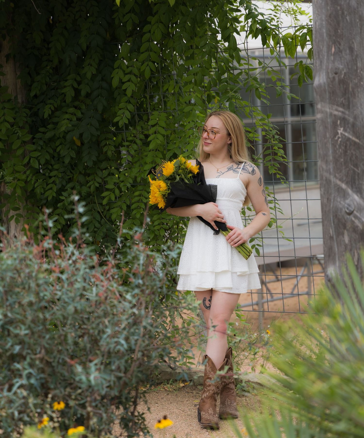 Blonde woman in white dress and cowboy boots holding sunflowers among lush greenery.