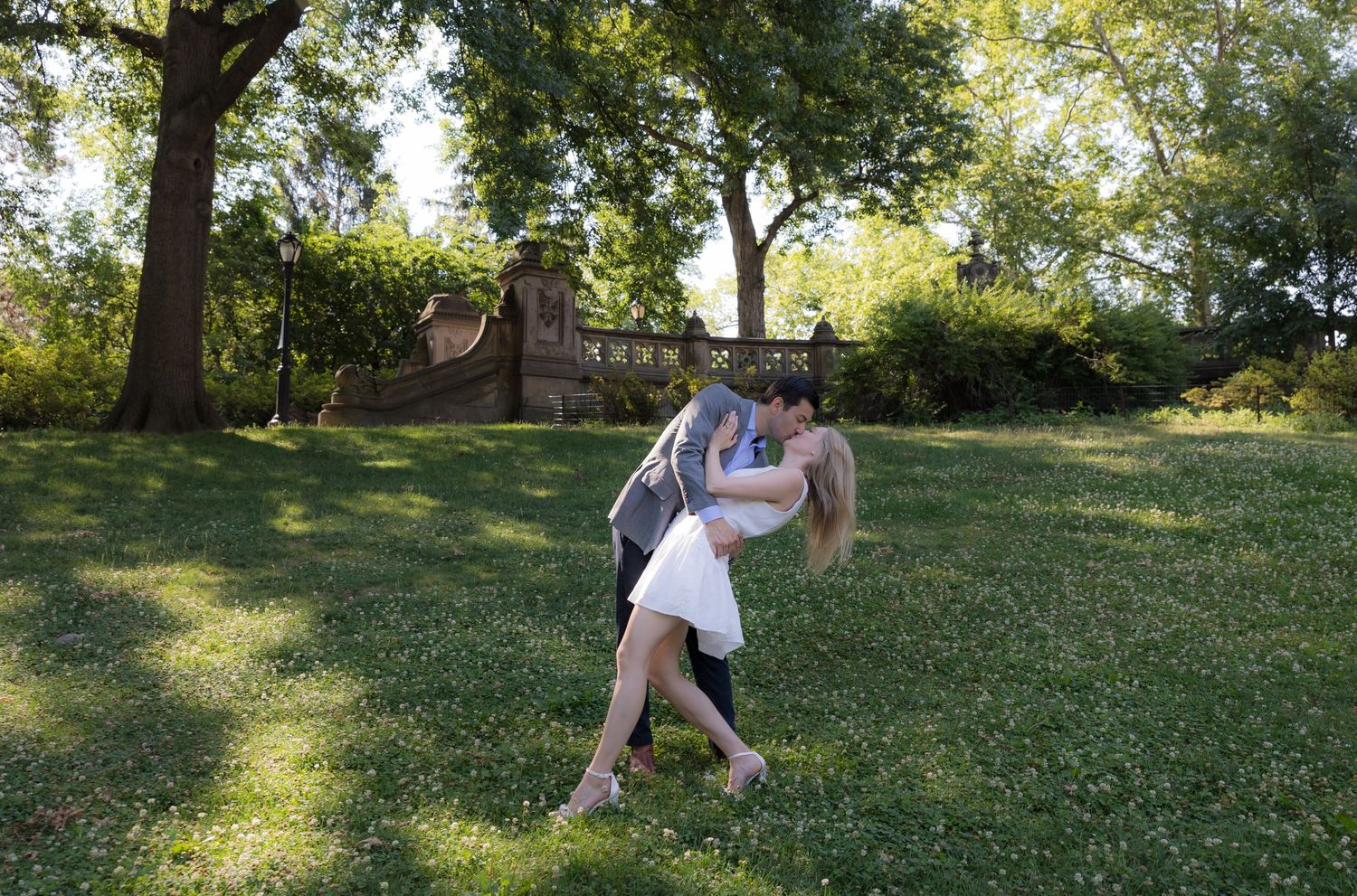 Romantic couple sharing a dip kiss on lush green grass in a sunlit park with trees in the background.
